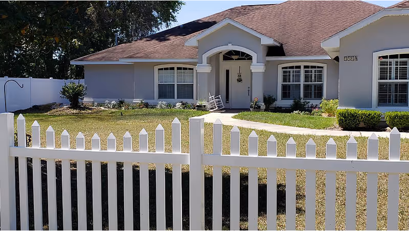 Front exterior view of a single-story house with a brown roof, light gray walls, white framed windows, and a white picket fence in the foreground. There is a small porch with a rocking chair and a walkway leading to the front door, surrounded by a lawn and some shrubs.