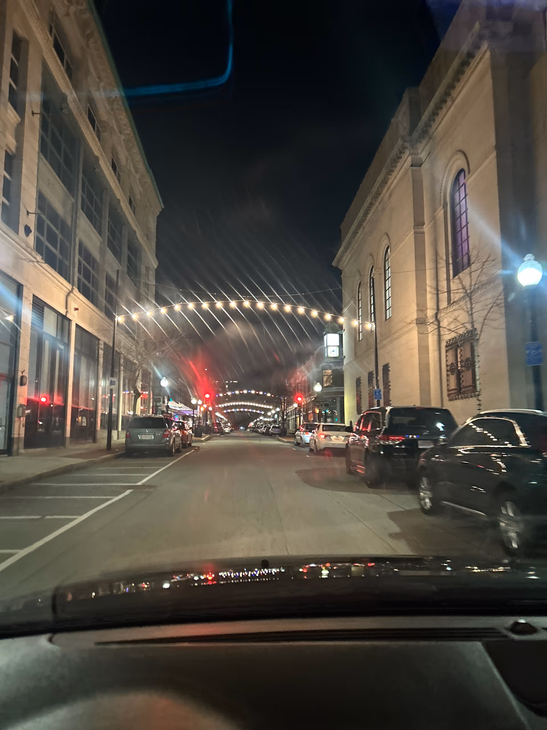 Nighttime city street viewed from inside a car, string lights line the avenue with parked cars.
