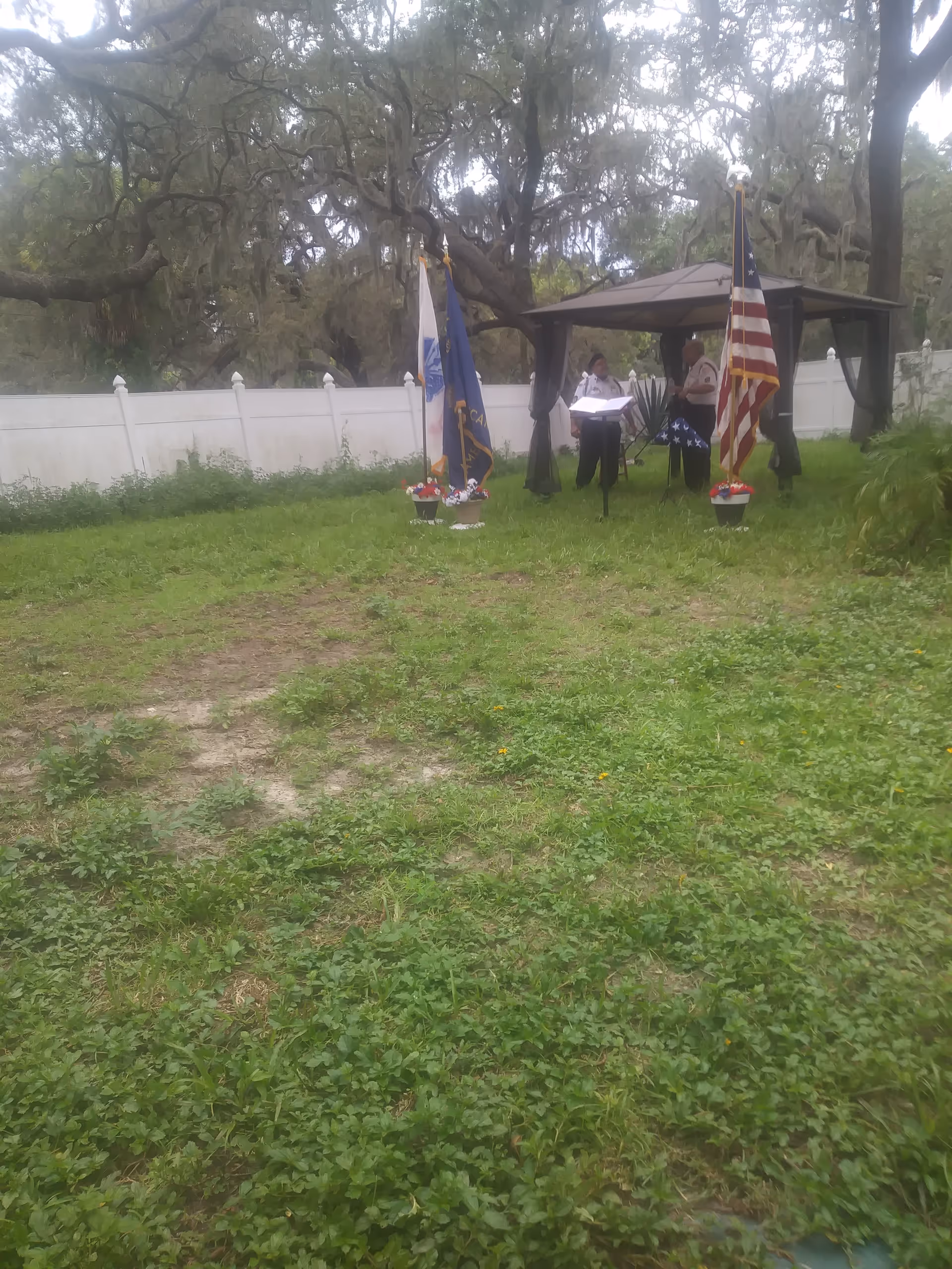 Outdoor ceremony at a grassy yard with gazebo, flags, podium, and attendees.