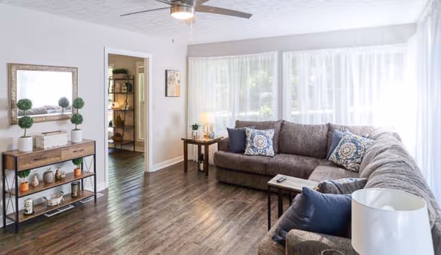 Sunlit living room with a large gray sectional sofa, wood floors, side tables, and a console table with decor beneath a wall mirror.