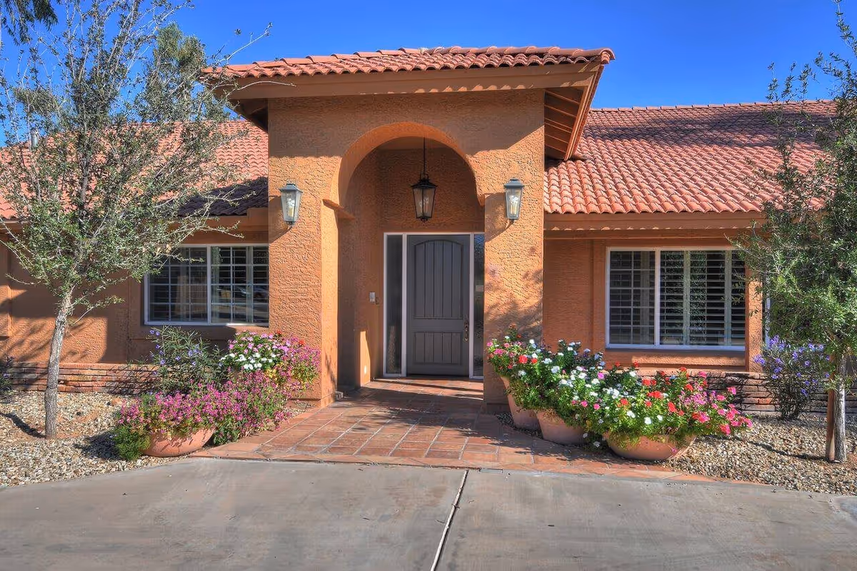 Front exterior view of a single-story building with a terracotta tiled roof and an arched entrance. The entrance has a dark wooden door, two wall-mounted lanterns, and a hanging lantern. There are flower pots with colorful flowers and small trees on either side of the entrance, and a clear blue sky in the background.