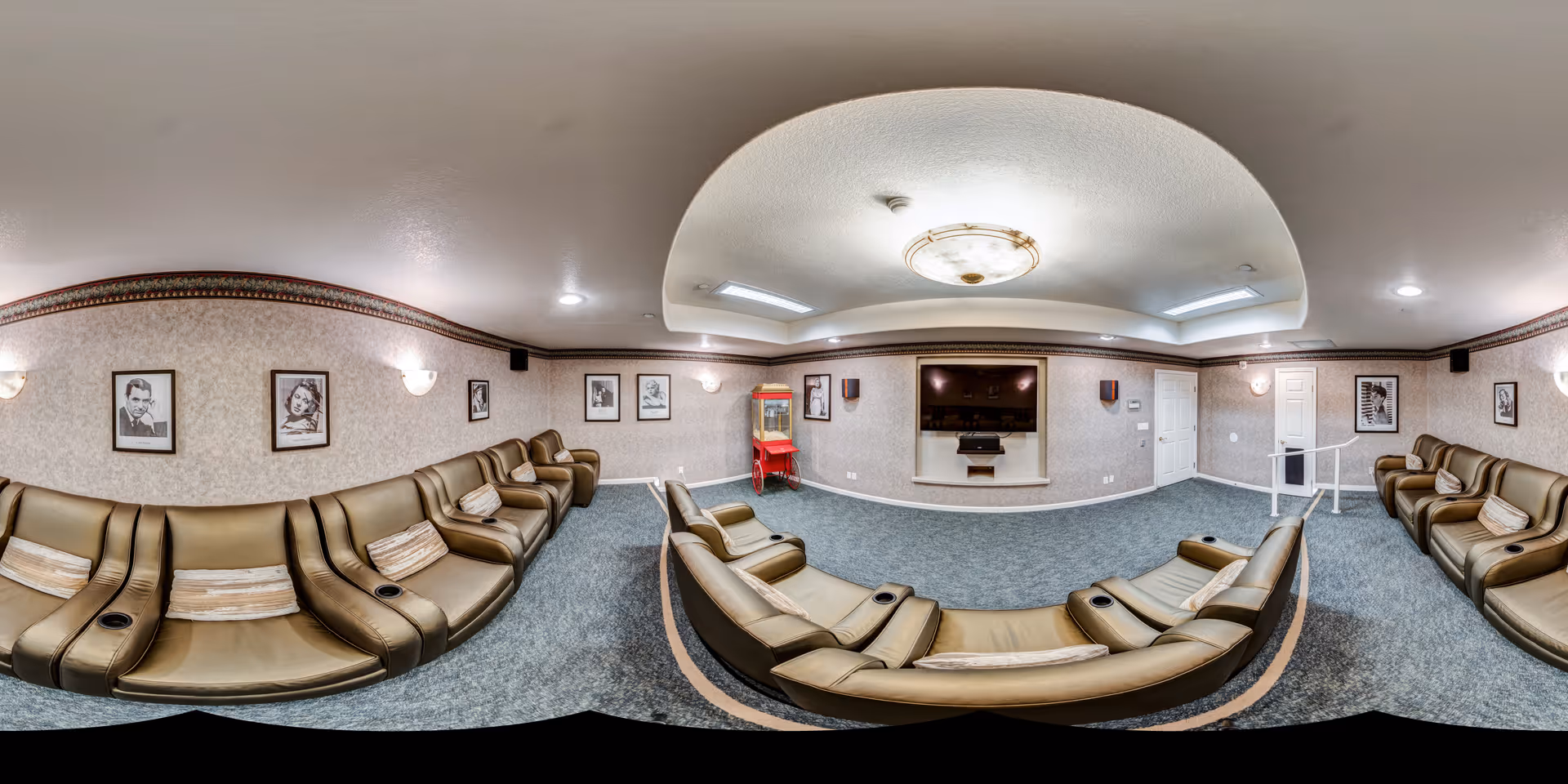A cozy entertainment room with curved rows of brown leather recliner chairs facing a large flat-screen TV mounted on a beige wall. The room has soft lighting from wall sconces and a ceiling fixture, framed black and white portraits on the walls, and a red popcorn machine in the corner. The carpet is blue, and the ceiling has a recessed dome design.