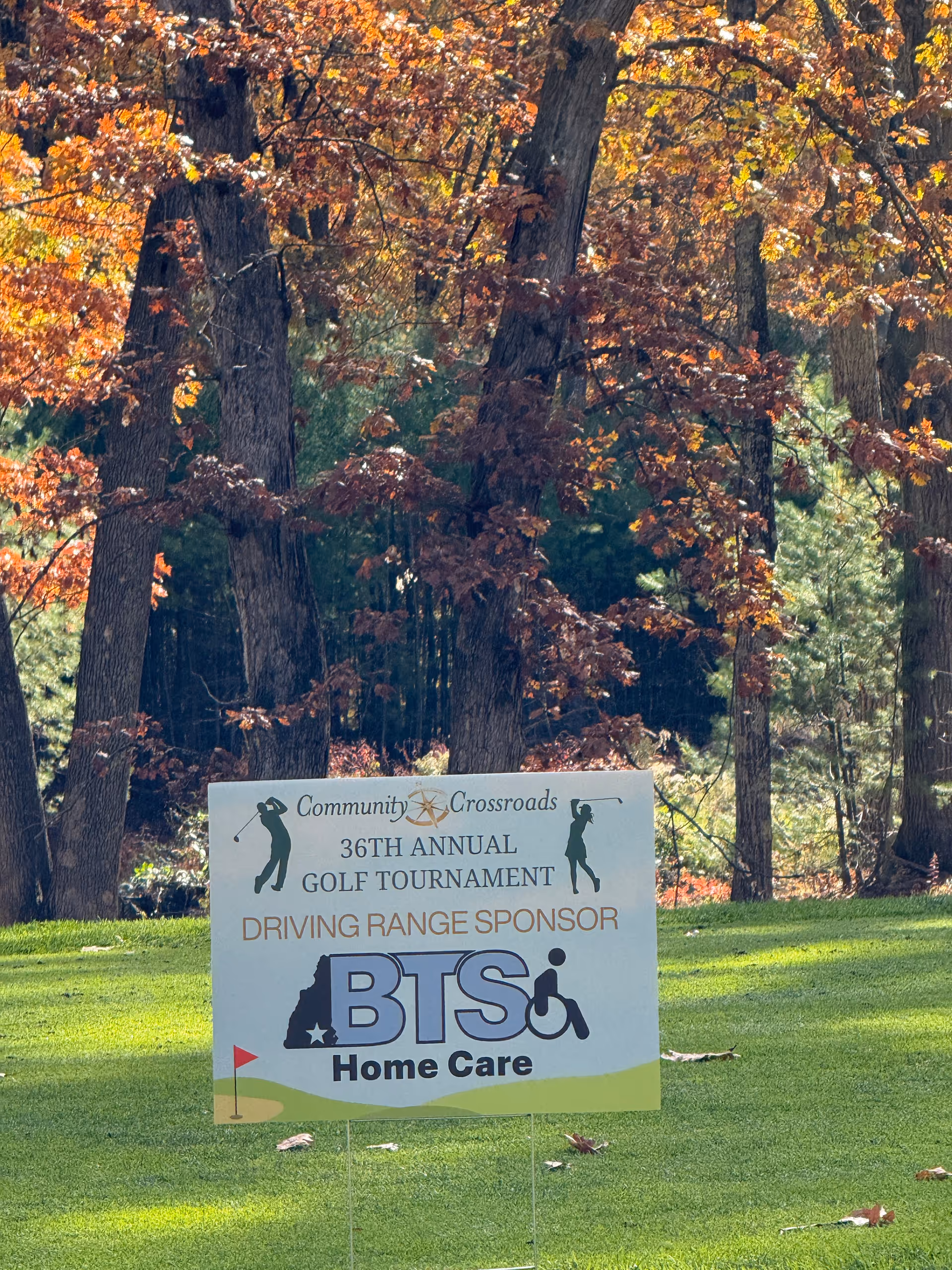 Outdoor scene with a BTS Home Care golf tournament sign among trees on a sunny course.