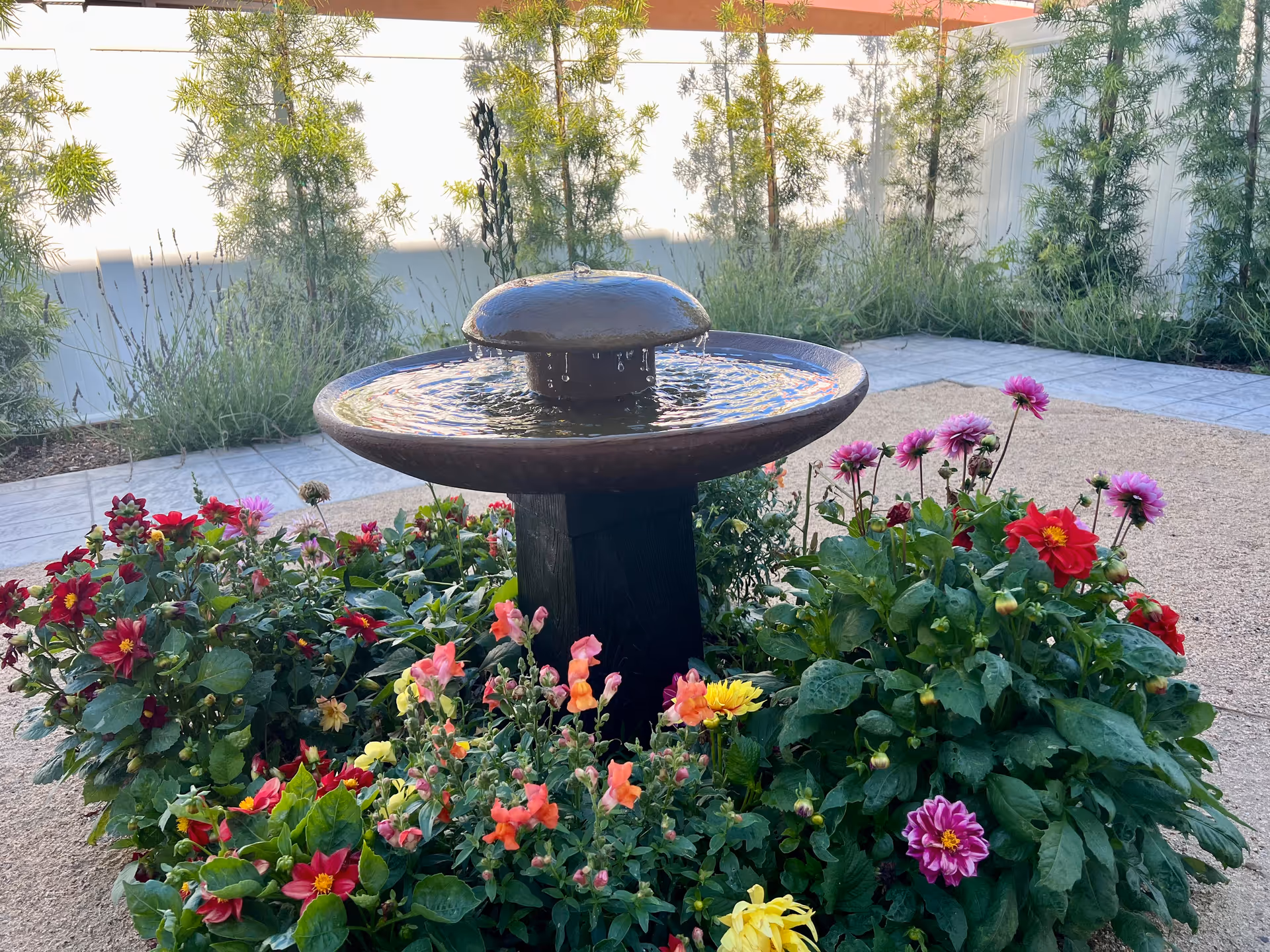 A round tiered fountain surrounded by vibrant flowers in a sunny outdoor courtyard.