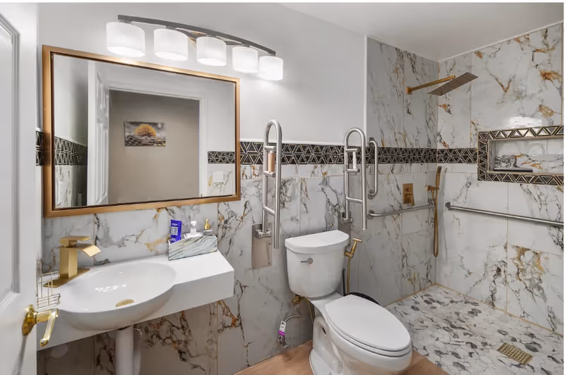 Marble-tiled bathroom with sink, mirror, toilet, and accessible shower with grab bars.