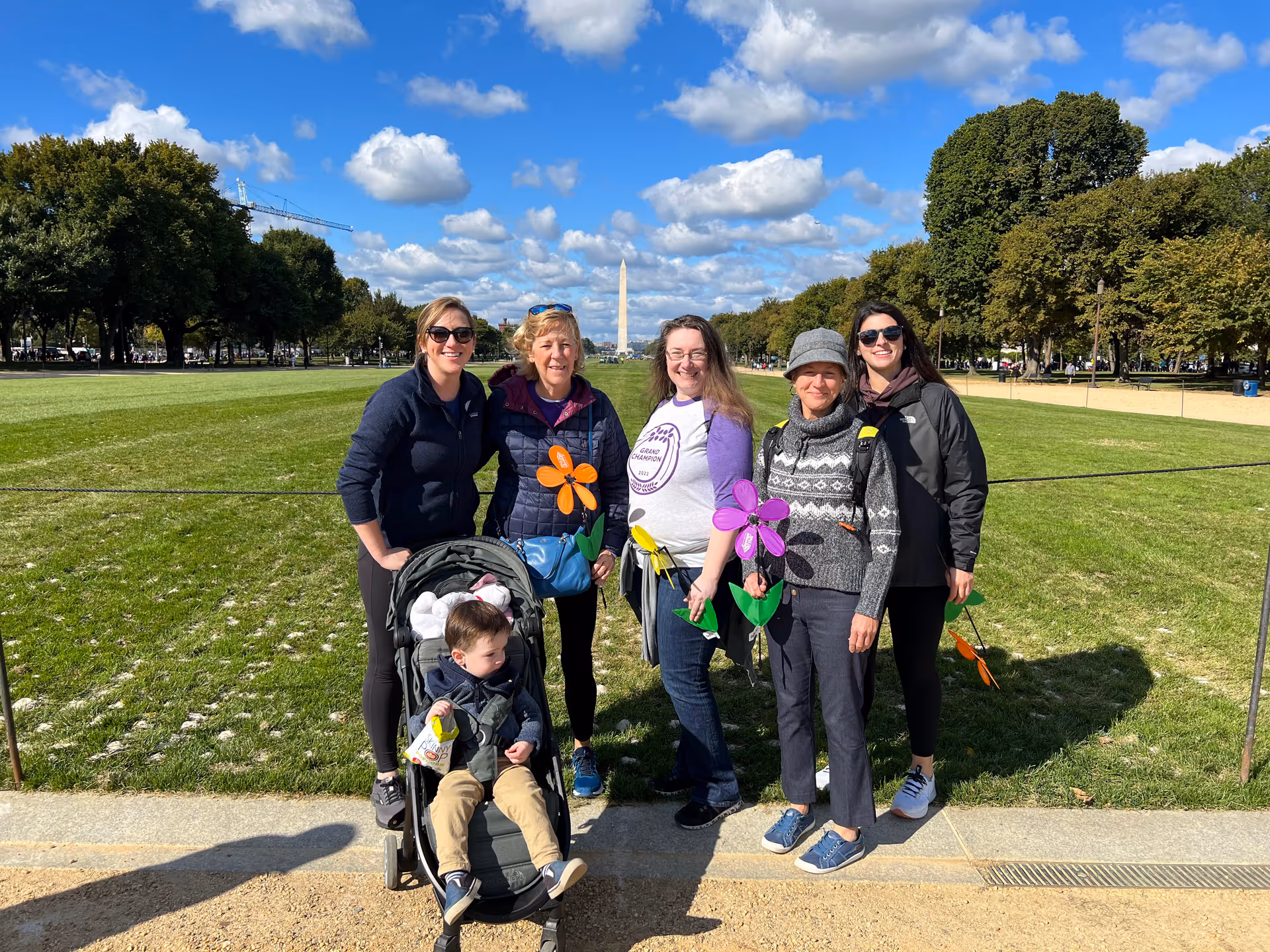 Group of five adults and a child in a stroller pose outdoors in a sunny park.
