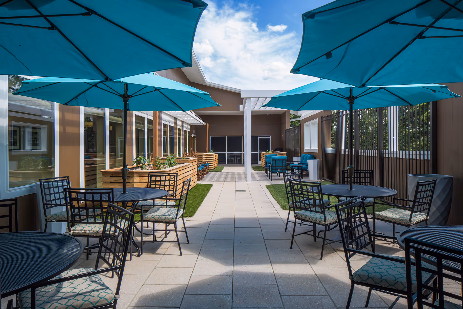 Outdoor courtyard with round metal tables, teal umbrellas, and chairs arranged along a central tiled walkway beside raised planters and seating.