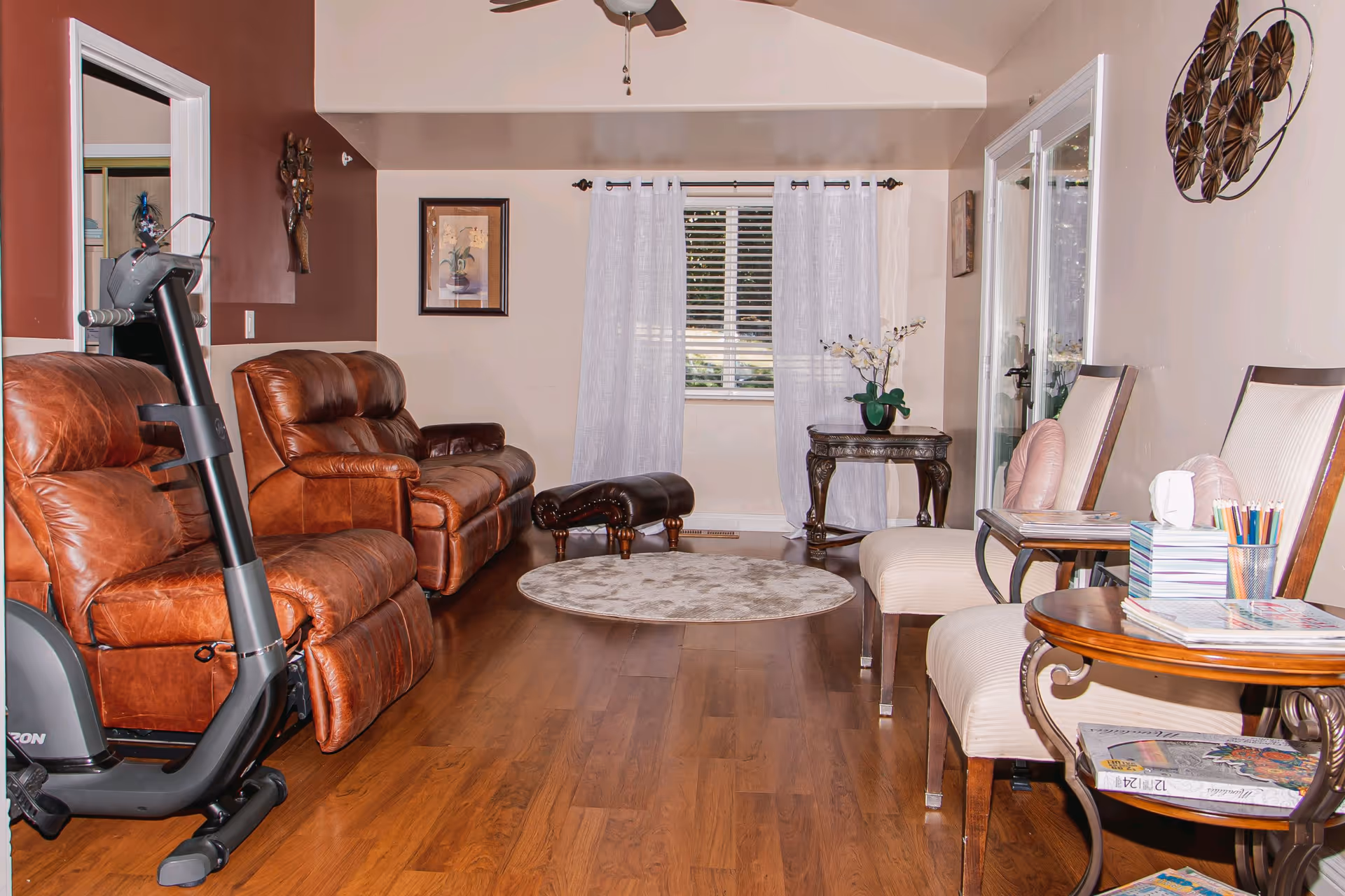 Cozy living room in a senior facility with brown leather seating, exercise bike, and window curtains.
