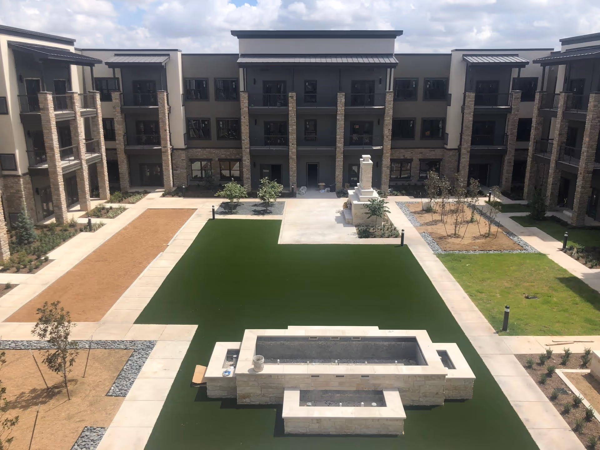 View of a three-story senior living facility courtyard with a central green lawn, walkways, a stone fountain, and surrounding balconies.