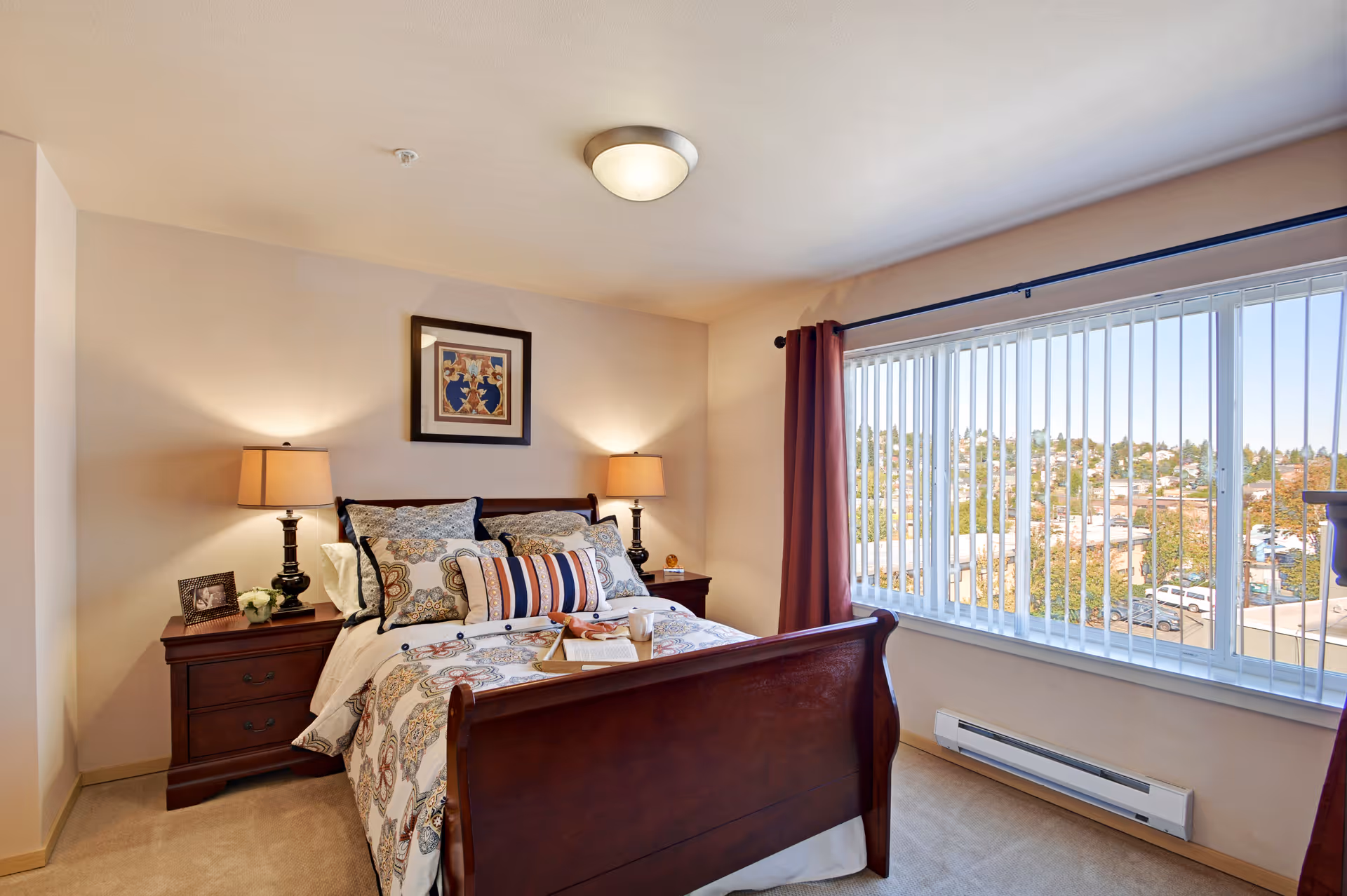 A cozy furnished bedroom in a senior living facility with a wooden bed and large window.