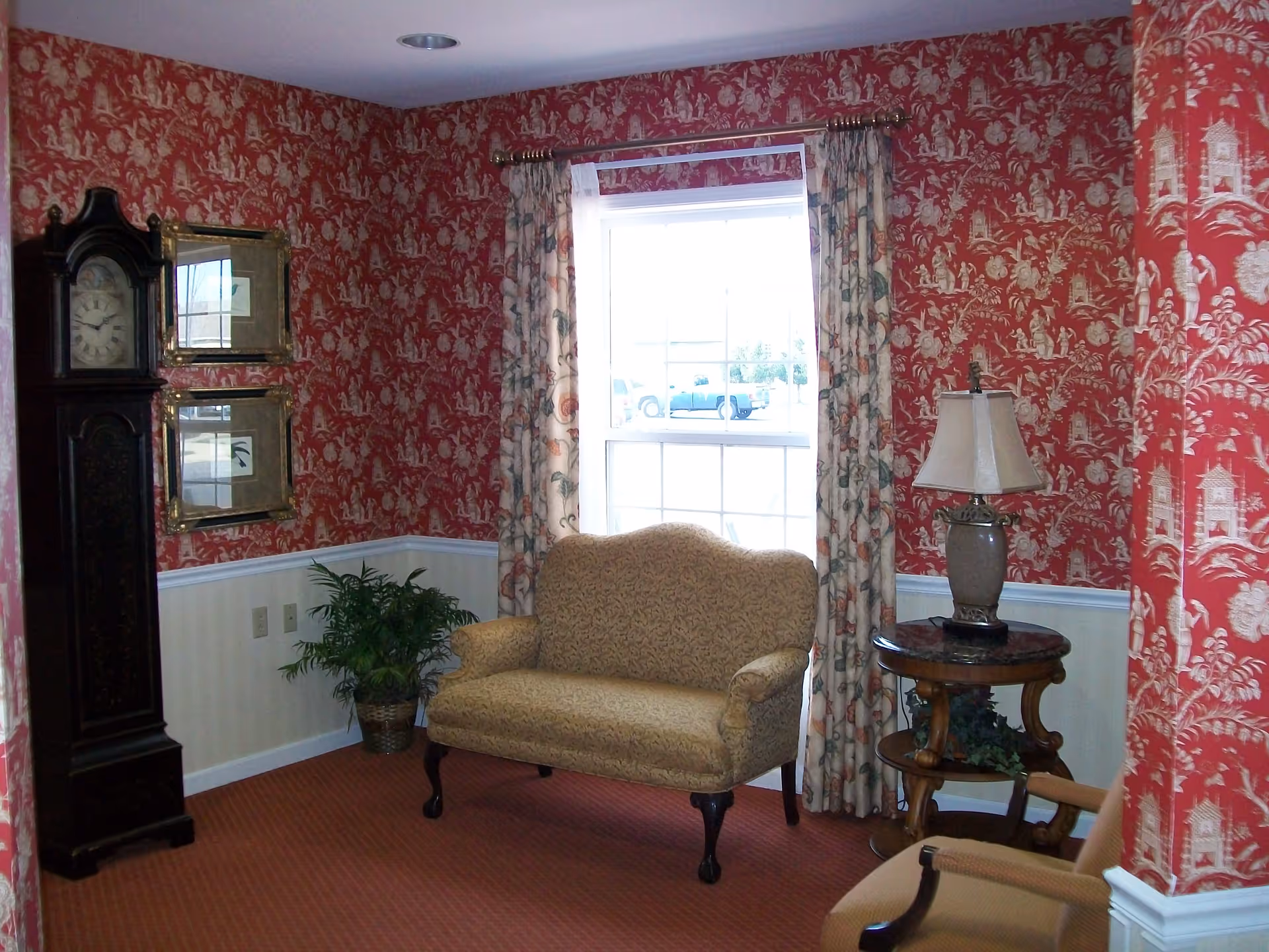 Cozy sitting area with red wallpaper, sofa, grandfather clock, and lamp by a window.