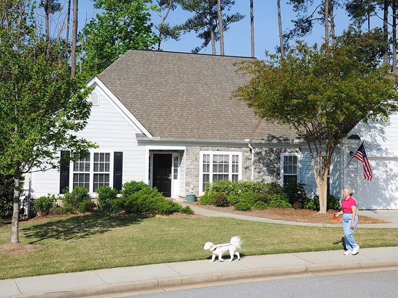 A white single-story house with a gray roof and black shutters surrounded by green bushes and trees. A woman wearing a pink shirt and blue jeans is walking a small white dog on a leash along the sidewalk in front of the house. An American flag is mounted near the garage.