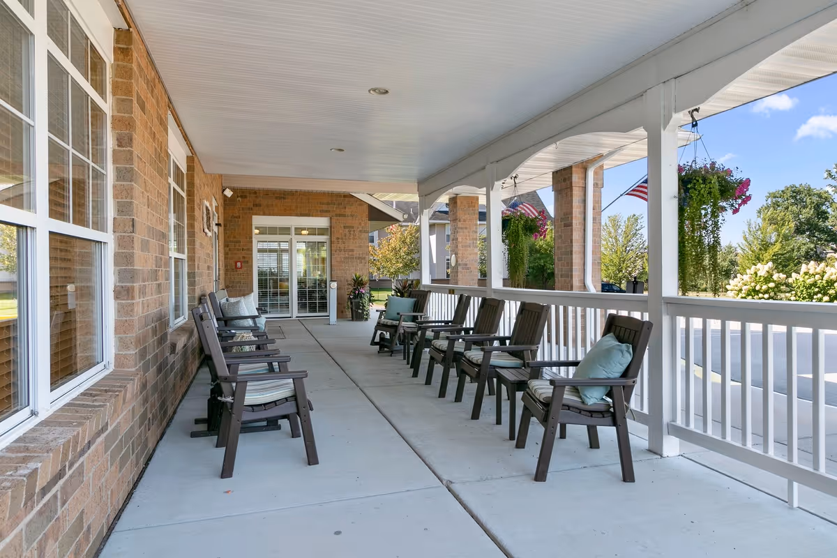 Covered front porch of a brick senior living building with a row of outdoor chairs and cushions along the entrance.