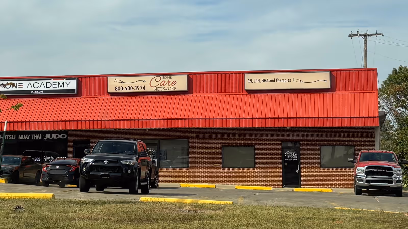 Red brick storefront with Home Care Network signs and several parked vehicles in front.