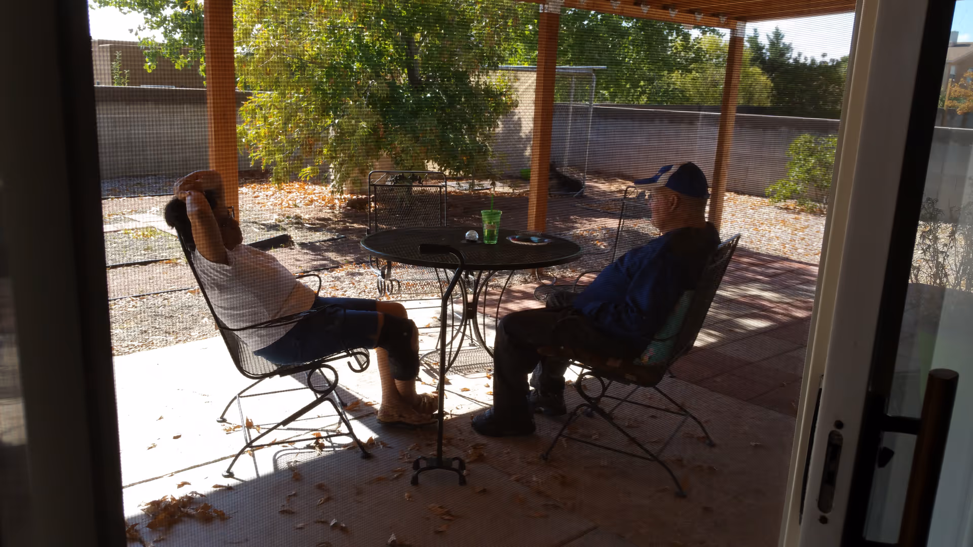 Two elderly residents sit at a round table on a shaded outdoor patio.
