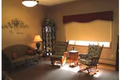 A cozy sitting area with a sofa, two upholstered rocking chairs, a side table, lamp, and a glass display cabinet in a warmly lit room.