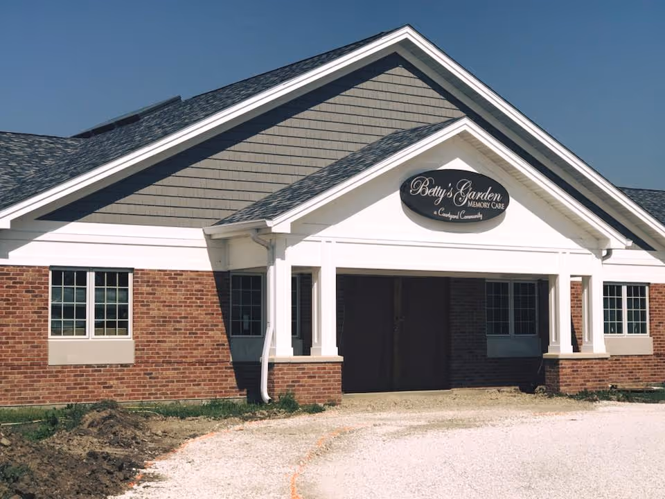 Exterior front view of a single-story brick building with white trim and a gray shingled roof under a clear blue sky. The building has a sign that reads 'Betty's Garden Memory Care a Crawford Community' above the entrance.