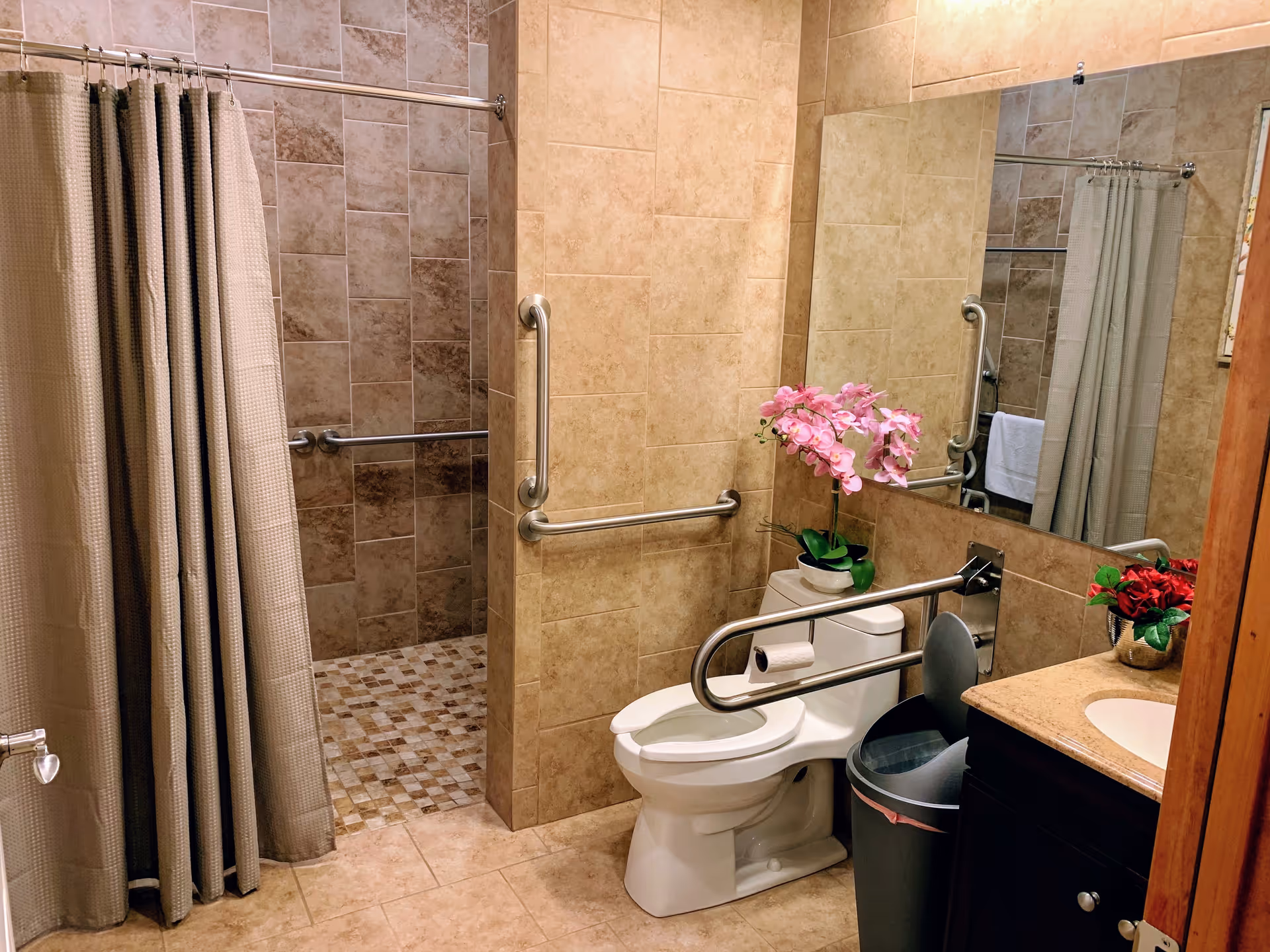 Accessible bathroom with a toilet and safety grab bars, a walk-in tiled shower with curtain, a vanity sink, and decorative flowers.