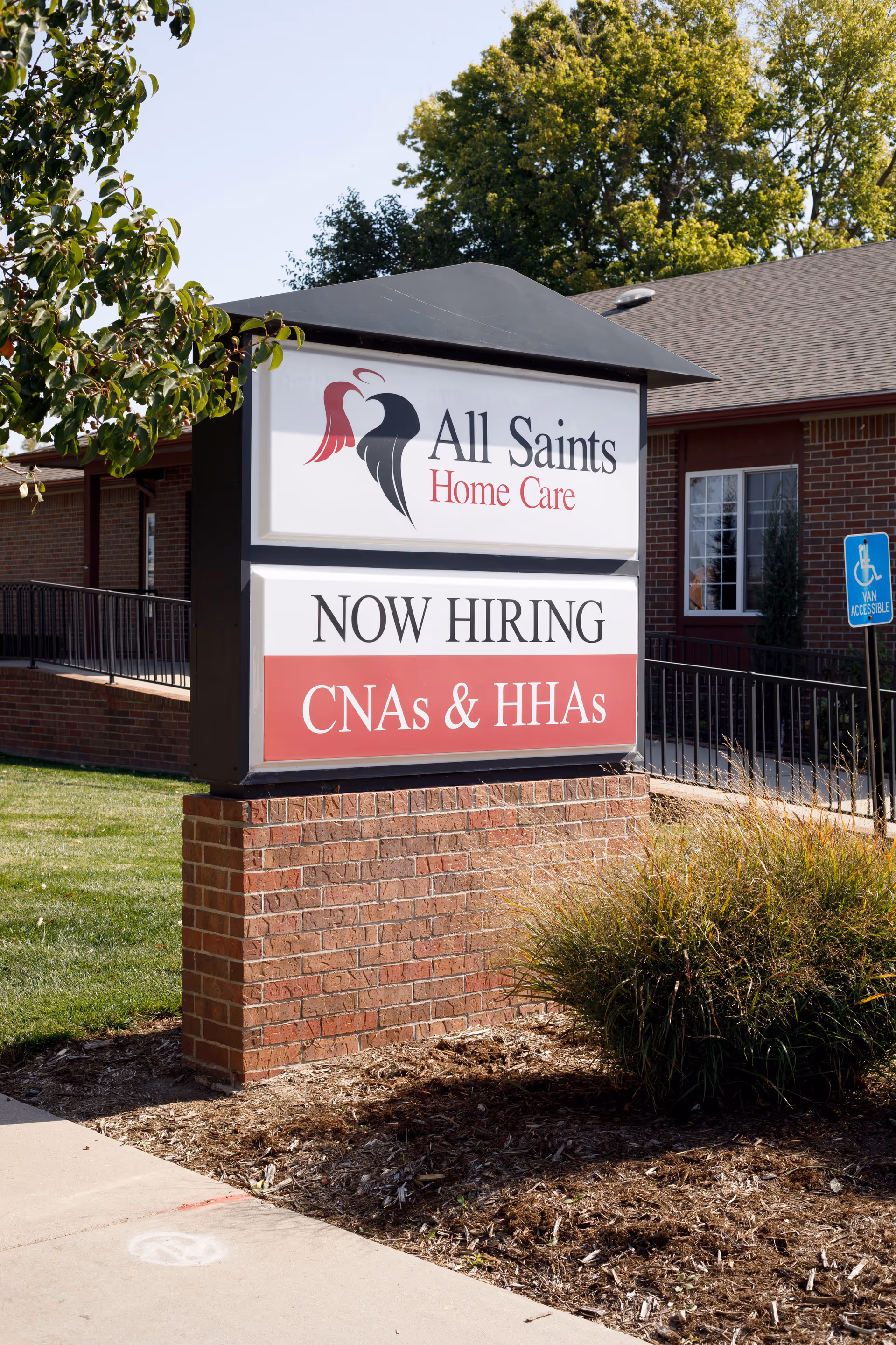 Exterior view of a brick facility sign for All Saints Home Care with Now Hiring