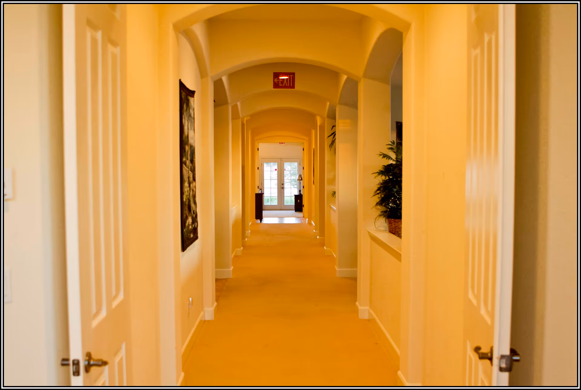 Long carpeted interior hallway with arched ceiling, doors on either side, plants and an exit door at the far end.