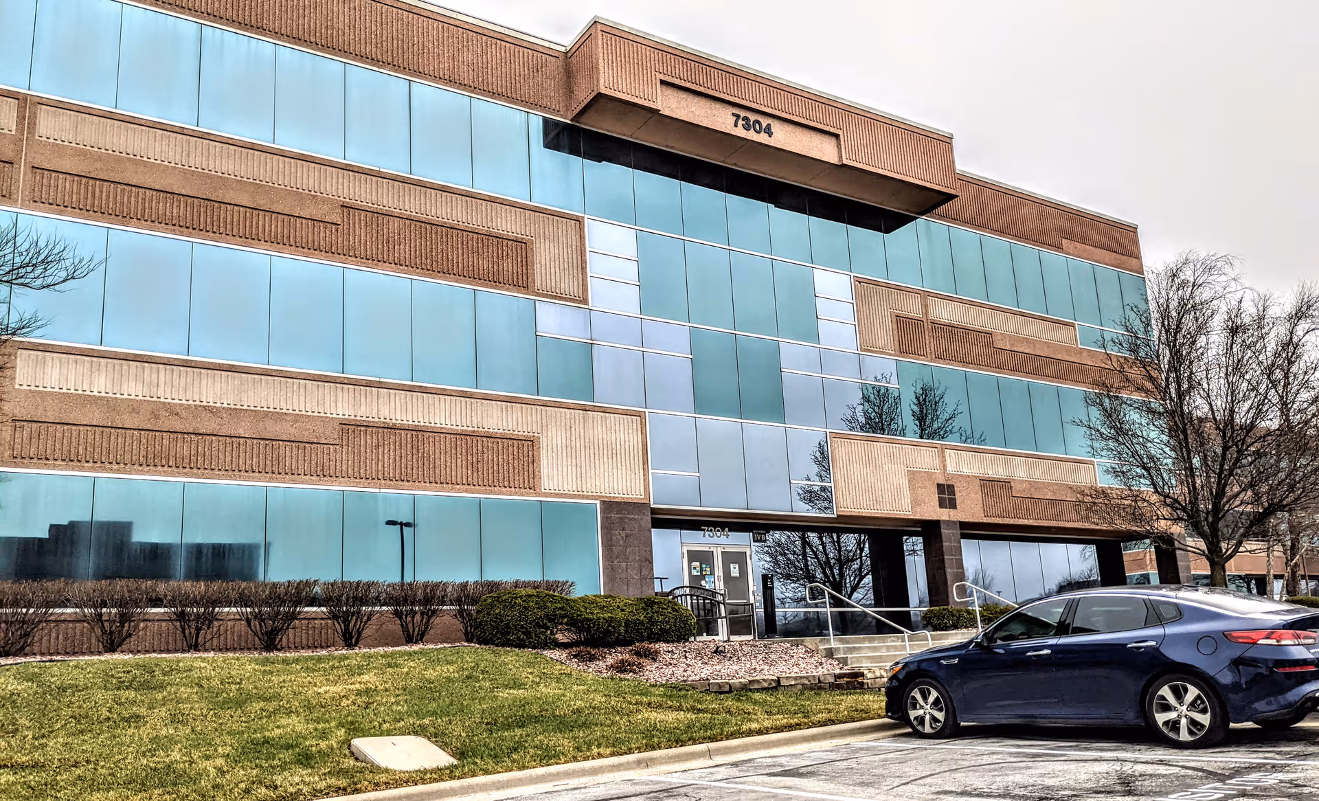 Modern glass-front office building with blue-tinted windows, landscaped entry, and parked car.