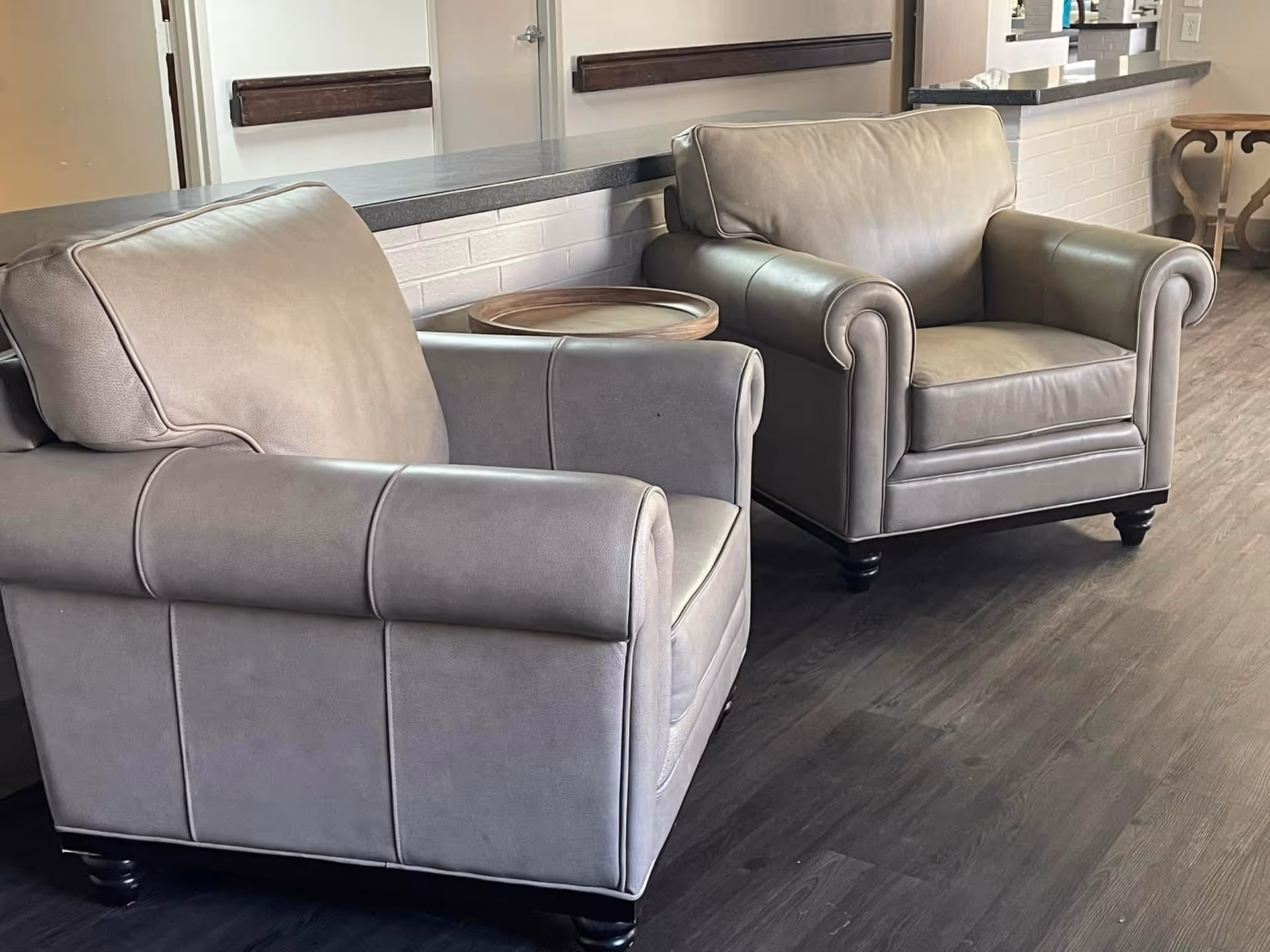 Two gray leather armchairs with a small round table between them sit on dark wood flooring in a facility common area.