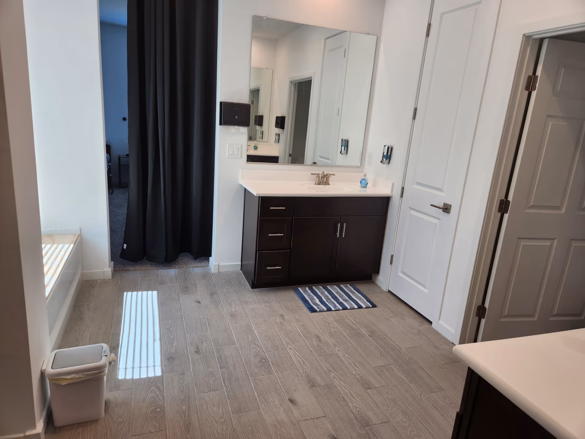 A clean and modern bathroom with light wood-look tile flooring, a dark wood vanity with a white countertop and sink, a large mirror above the sink, a striped blue and white rug on the floor, a white trash bin near a window with blinds, and white doors. A dark curtain separates the bathroom from an adjacent room.