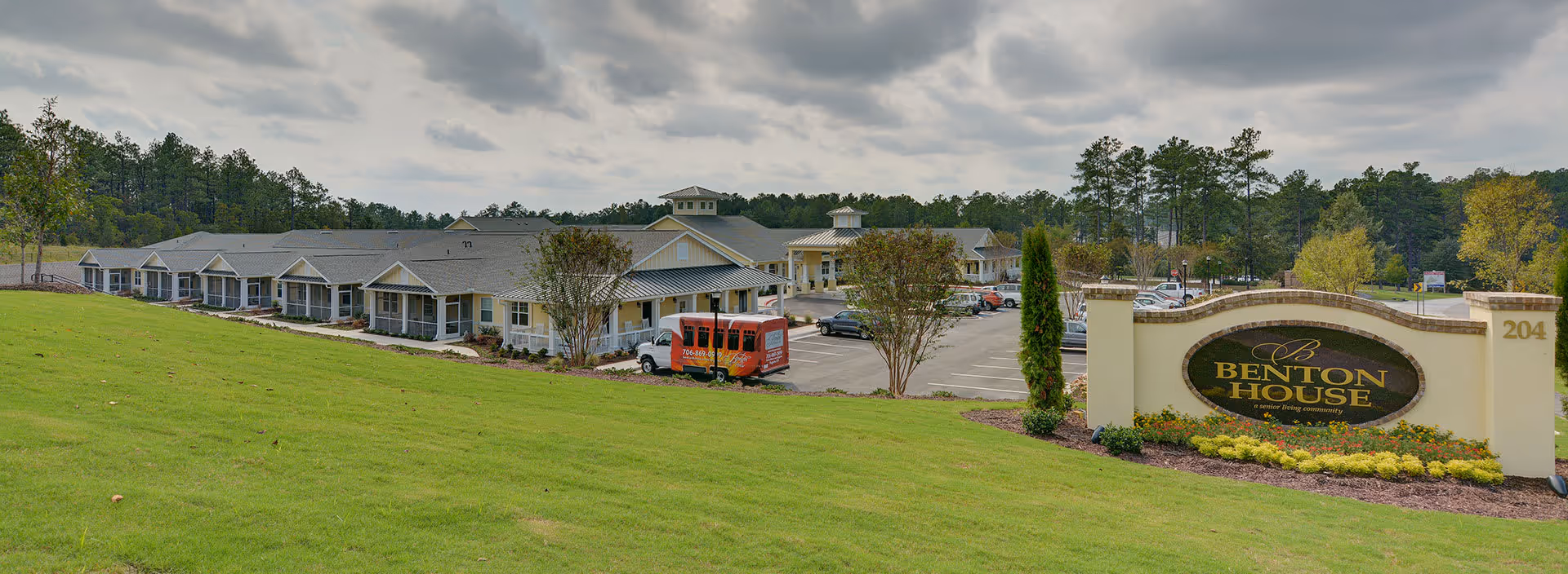 Exterior view of a senior living community named Benton House, showing a large single-story building with a parking lot and a green lawn in the foreground under a cloudy sky.