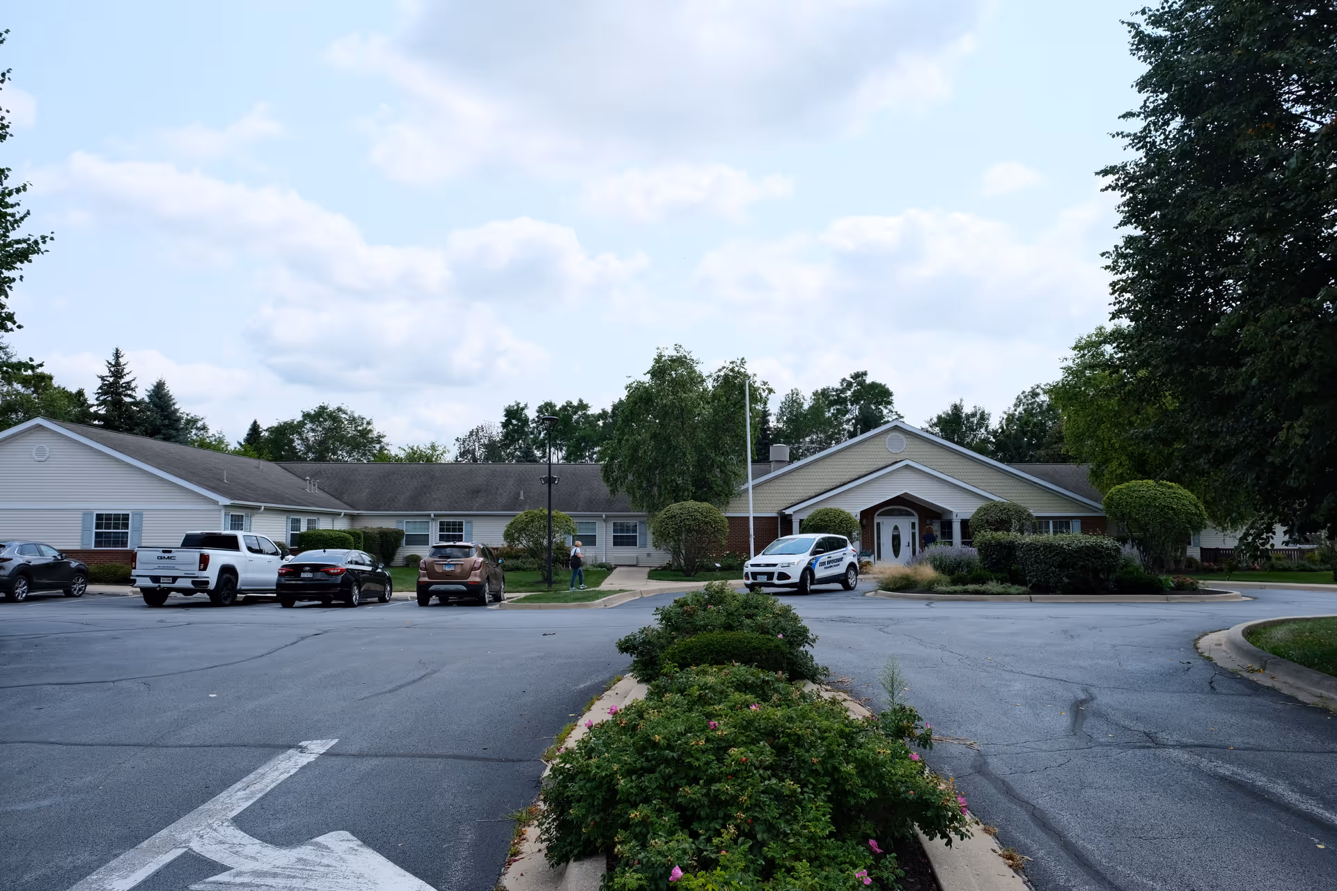 Front entrance of a single-story senior living facility with a driveway, parked cars, and landscaped greenery.