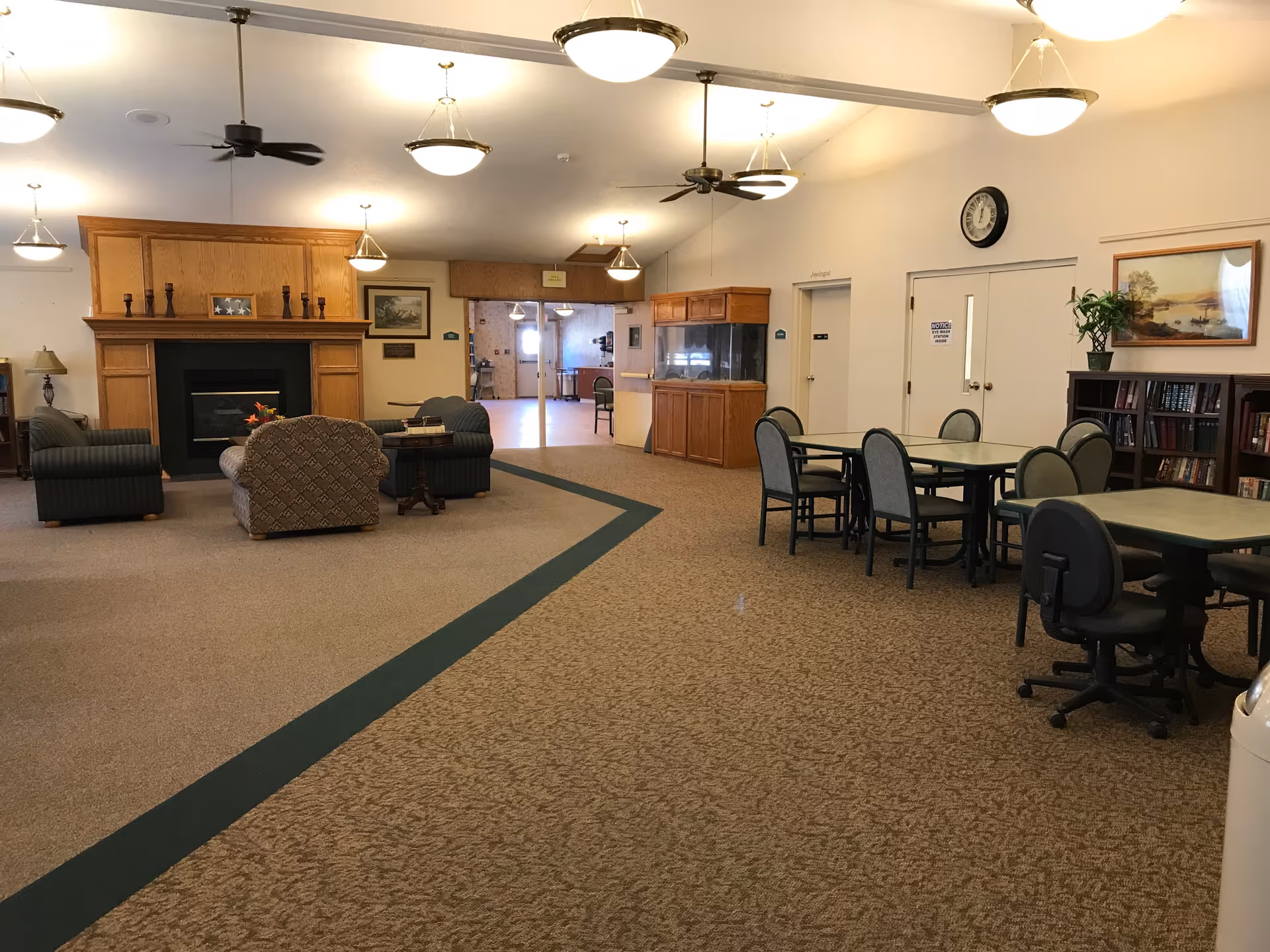 Interior view of a senior living facility common area with seating arrangements including armchairs and tables with chairs. There is a fireplace with a wooden mantel, ceiling fans, hanging lights, a clock on the wall, bookshelves, and a large fish tank. The space is carpeted and well-lit.