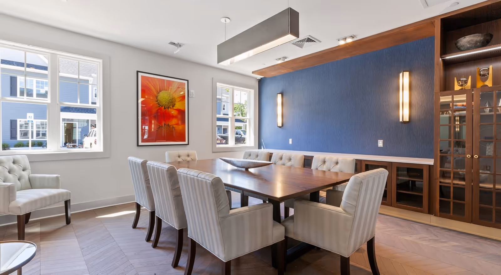 A modern dining room with a large wooden table surrounded by eight upholstered chairs. The room features two large windows letting in natural light, a vibrant orange flower artwork on the wall, a blue accent wall with two vertical wall lights, and built-in wooden cabinetry with glass doors.
