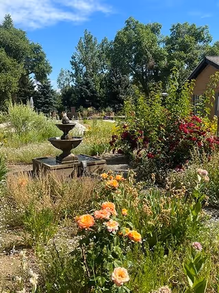 Sunny garden with blooming flowers, a tiered stone fountain, and trees beside a building.