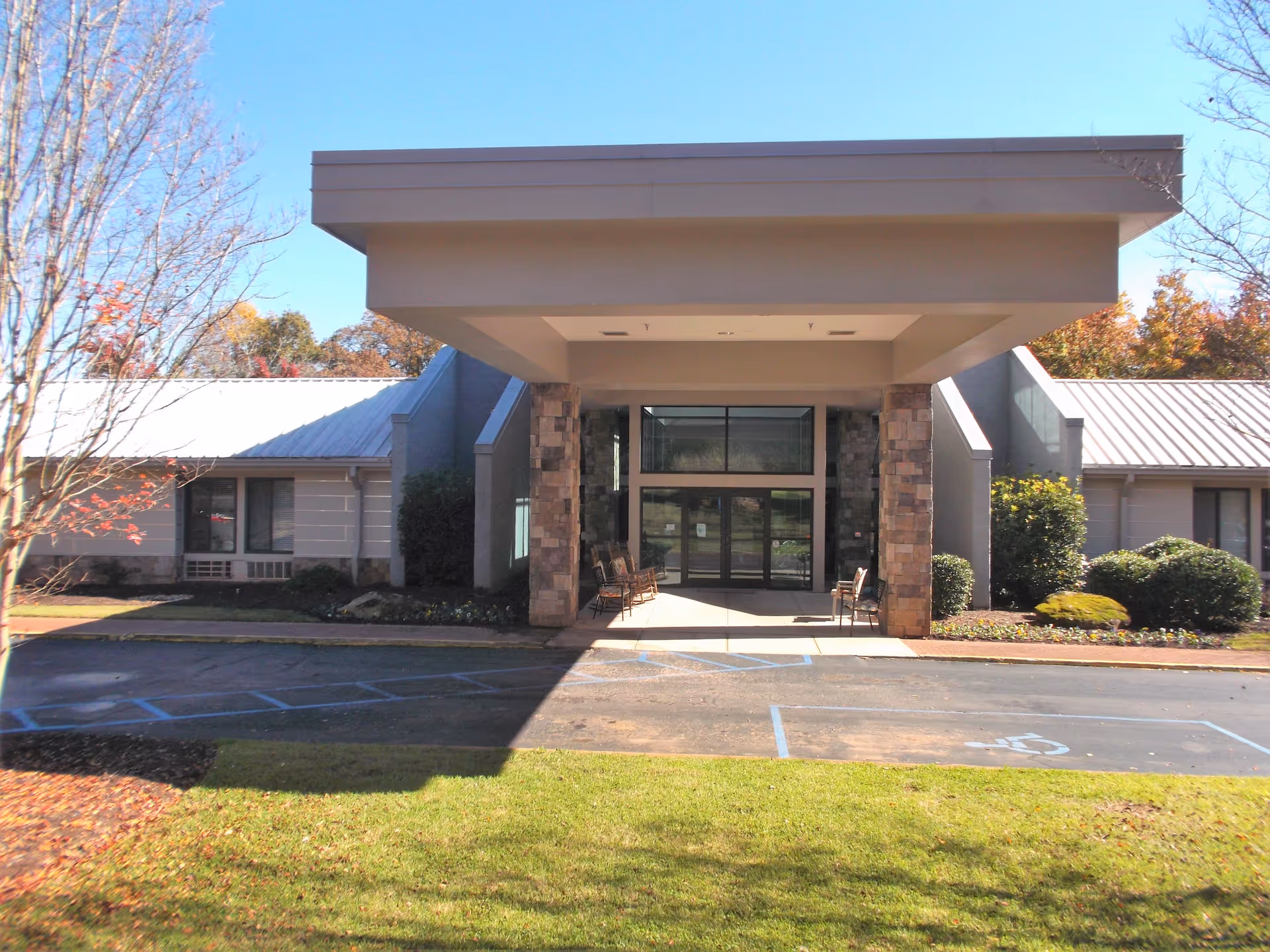 Front exterior view of TerraBella Summit facility showing a covered entrance with stone pillars, glass doors, and surrounding landscaping including bushes and trees with autumn foliage.