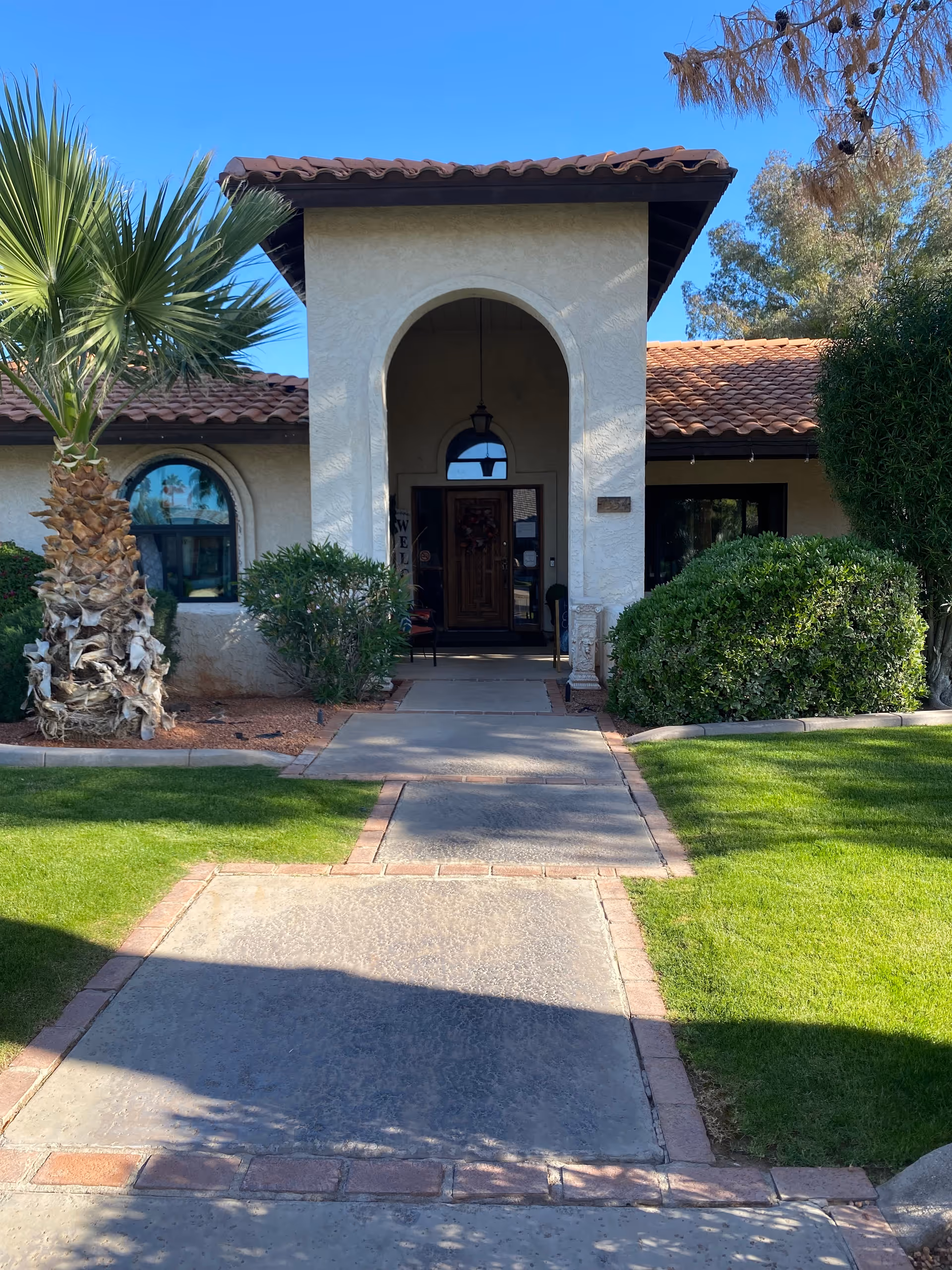 Front entrance of a single-story stucco building with a tiled roof, arched doorway, walkway, palm tree and landscaping.