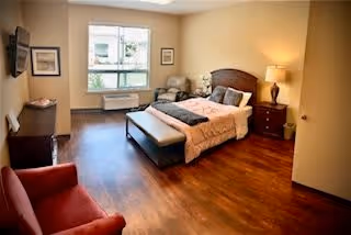 Furnished bedroom with a bed, bench at the foot, armchairs, a nightstand with lamp, a dresser with TV, and a window.