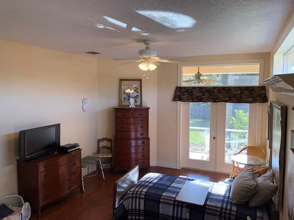 A bedroom with a bed covered in a blue and white plaid blanket, a small table on the bed, a wooden dresser with a TV on top, a tall chest of drawers, a chair, and glass doors leading to an outdoor area with greenery. The room has a ceiling fan with lights and wooden flooring.