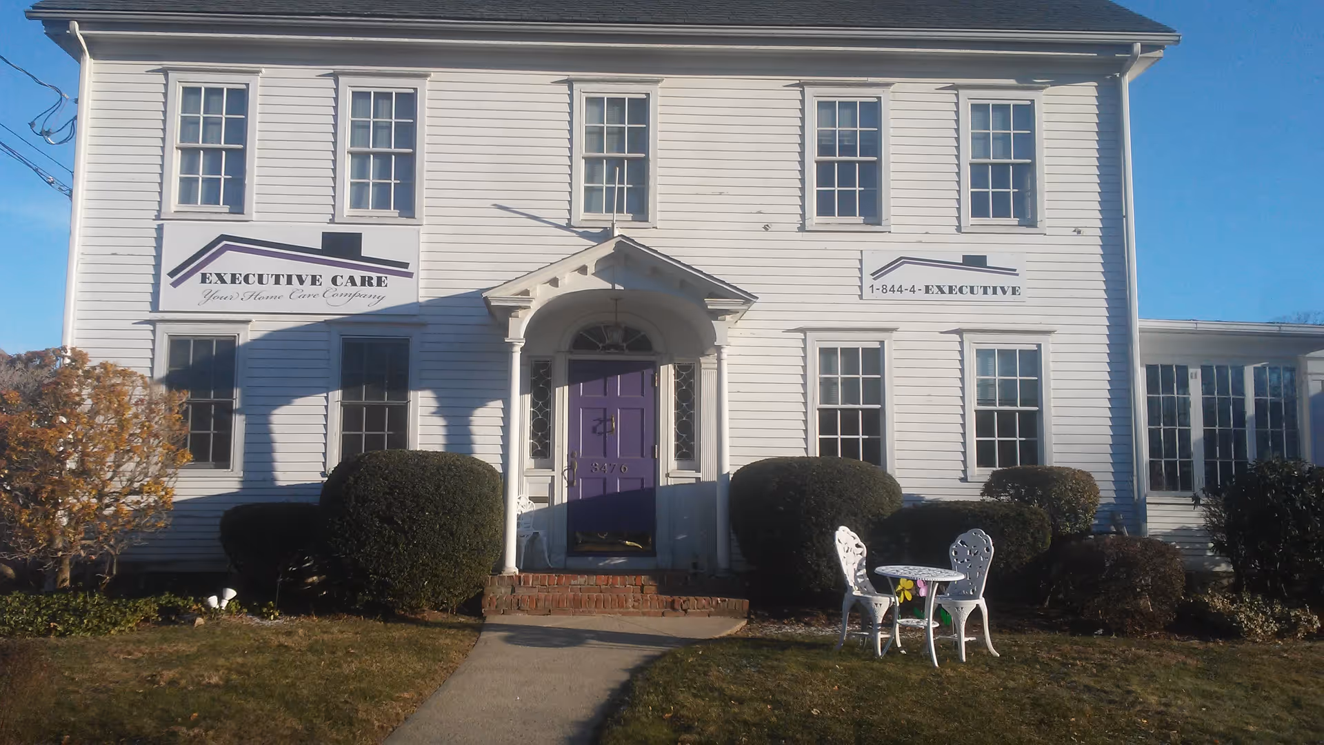 White two-story senior facility with purple front door and small outdoor seating near shrubs