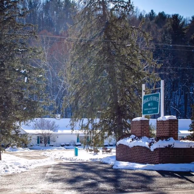 Snow-covered entrance to Highland House by American Retirement Homes with a brick sign and tall evergreen trees, set against a backdrop of a wooded hillside.