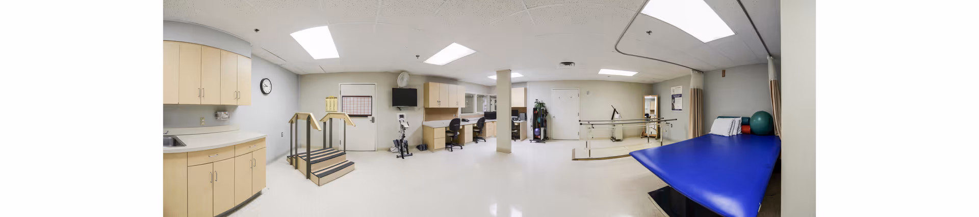 Panoramic view of a physical therapy room with parallel bars, a blue treatment table, practice stairs, exercise equipment, cabinets and workstations.