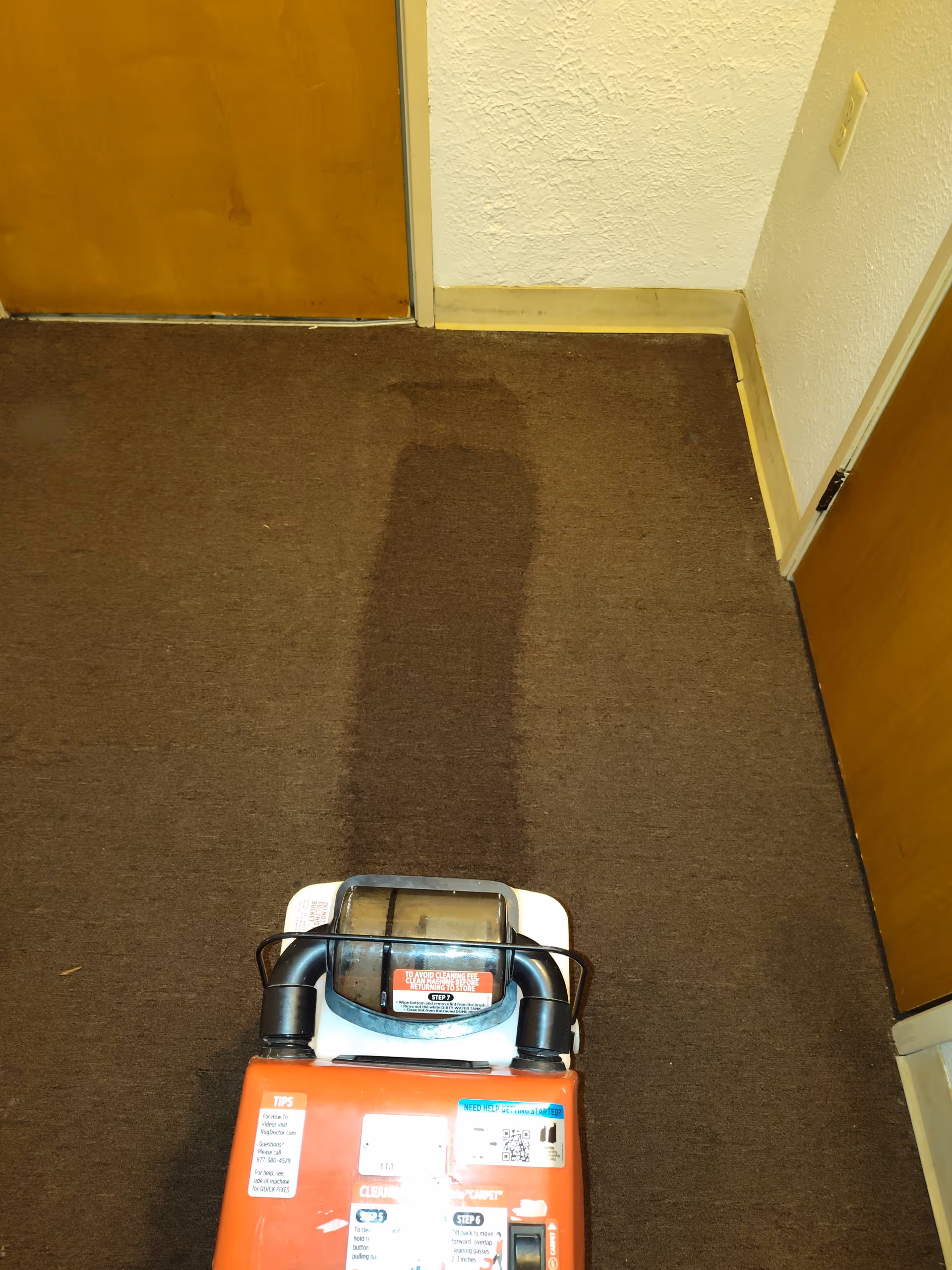 Brown carpet hallway with a red cleaning machine near a closed wooden door in a facility