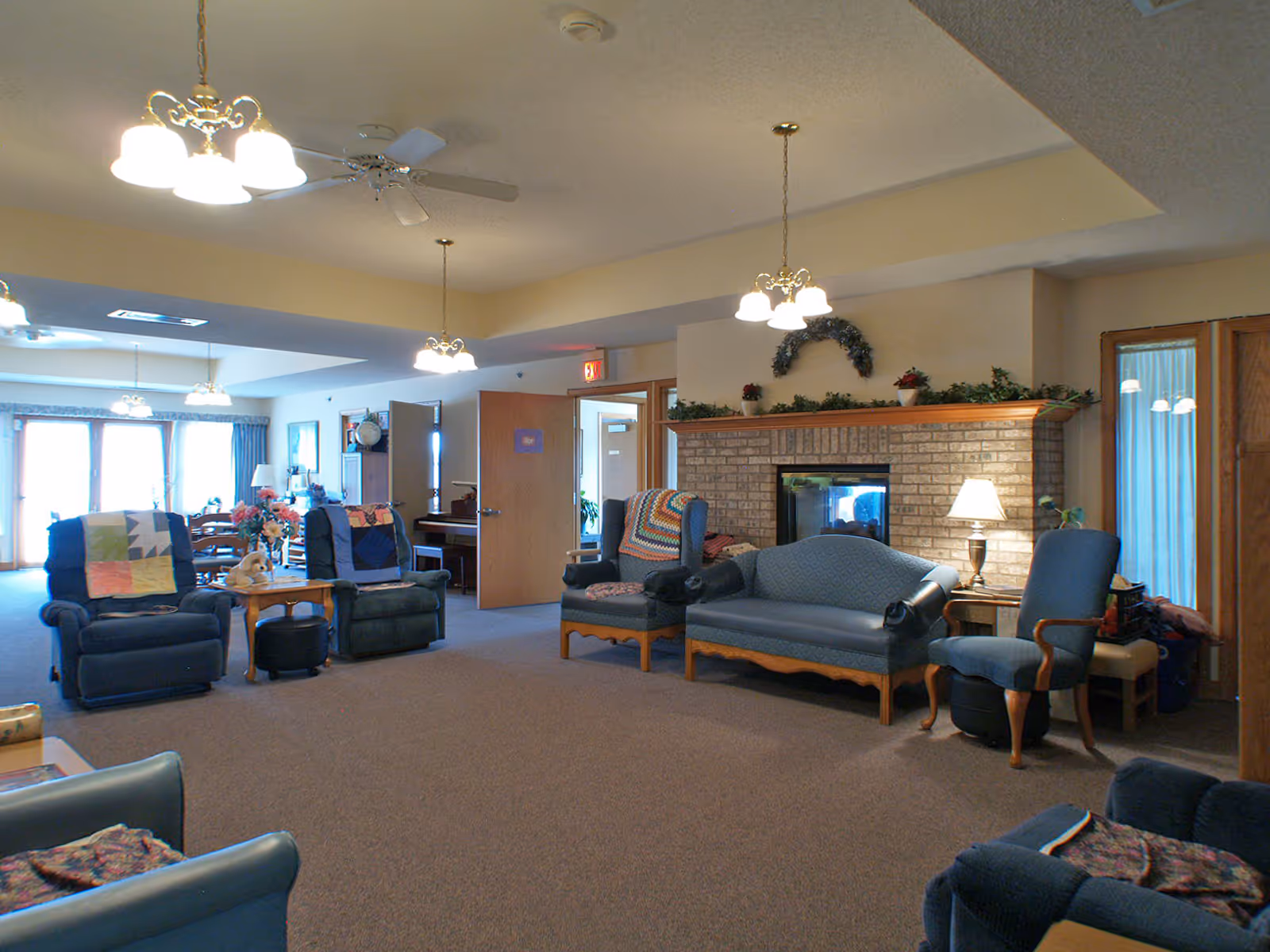 A cozy living room area in a senior living facility with multiple upholstered chairs and a loveseat arranged around a brick fireplace. The room is softly lit by ceiling lights and table lamps, with decorative plants and a wreath above the fireplace. In the background, there is a dining area with a table and chairs near large windows with curtains.