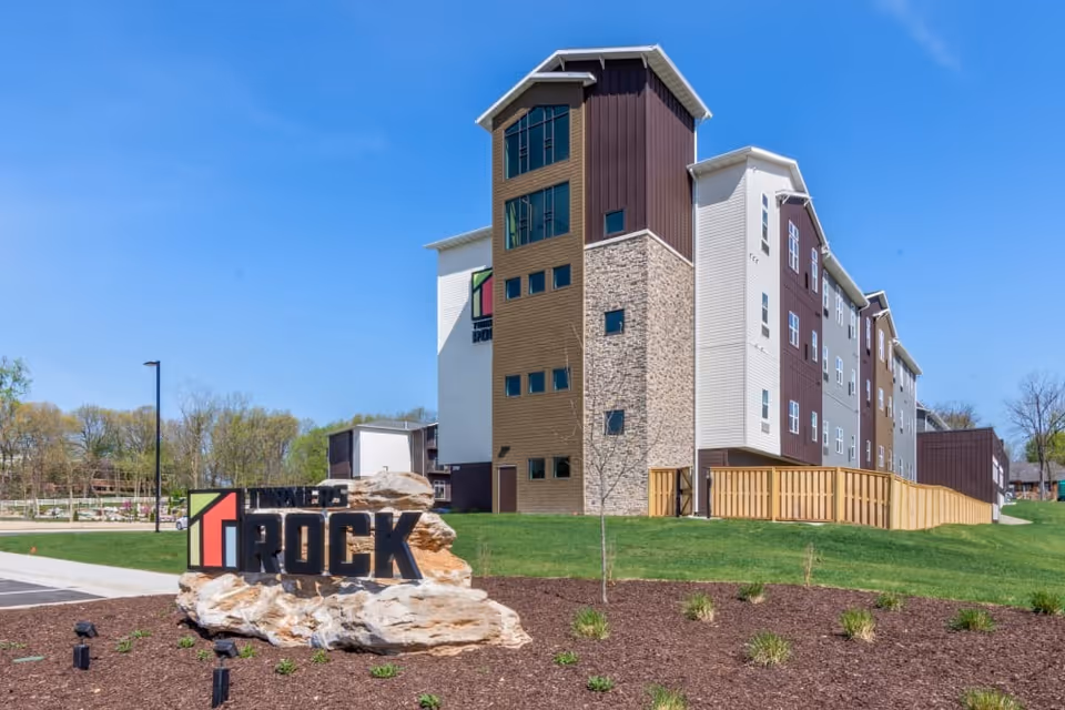 Exterior view of a multi-story senior living facility building with a mix of brown, beige, and white siding, stone accents, and multiple windows under a clear blue sky. In the foreground, there is a landscaped area with mulch, small plants, and a large rock featuring a sign that reads 'Turners Rock'.