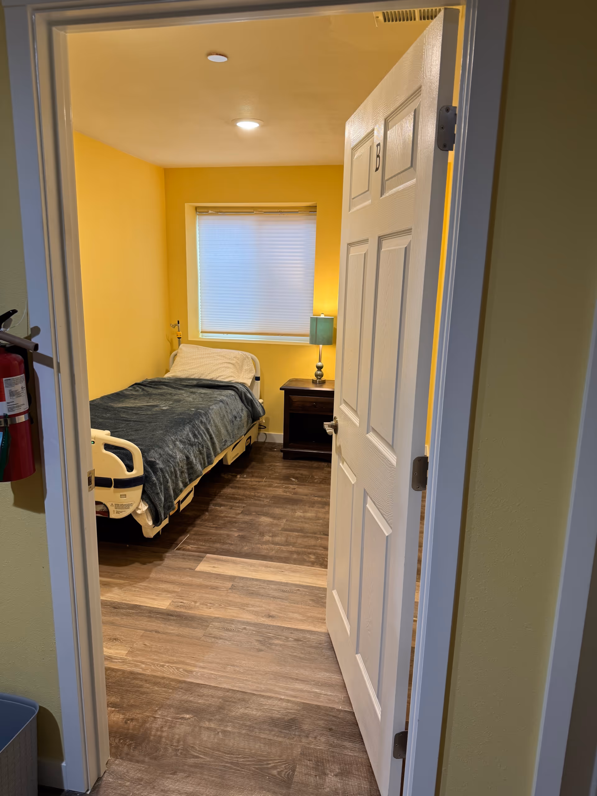 Yellow walls bedroom with a single bed, nightstand, lamp, and window in a senior living facility.