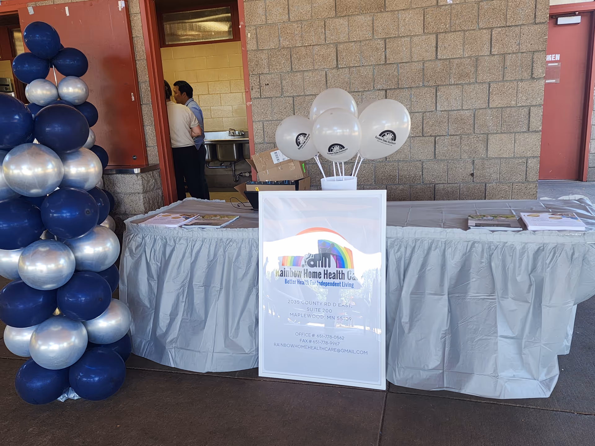 Outdoor display table with Rainbow Home Health Care sign, balloons, and staff near doorway.