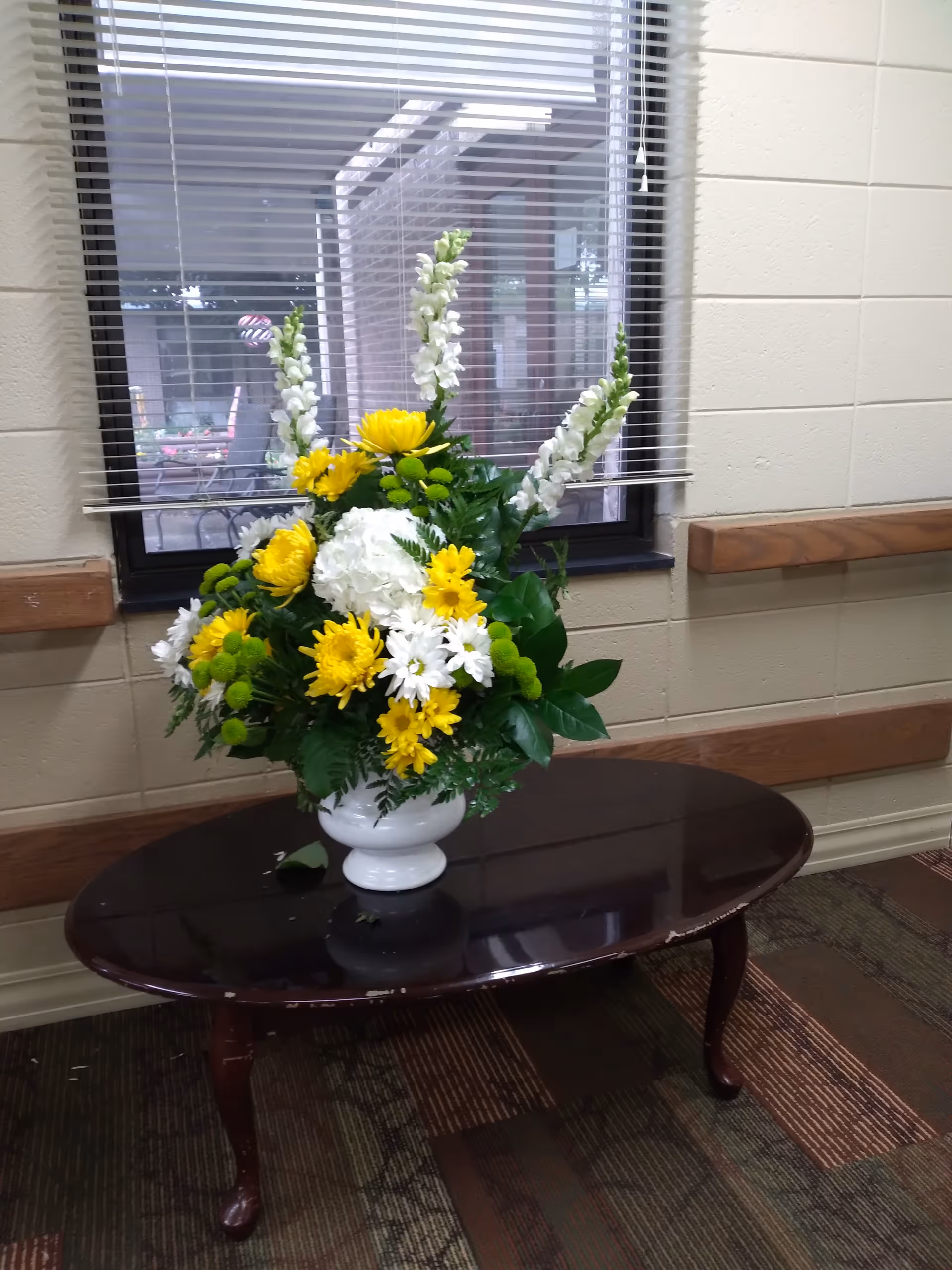 Yellow and white floral arrangement in a white vase on a wood table by a window.