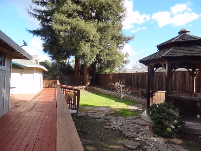 Outdoor area with a wooden deck on the left, a grassy lawn in the center, a stone pathway, and a wooden gazebo on the right. Tall trees and a wooden fence are visible in the background under a partly cloudy sky.