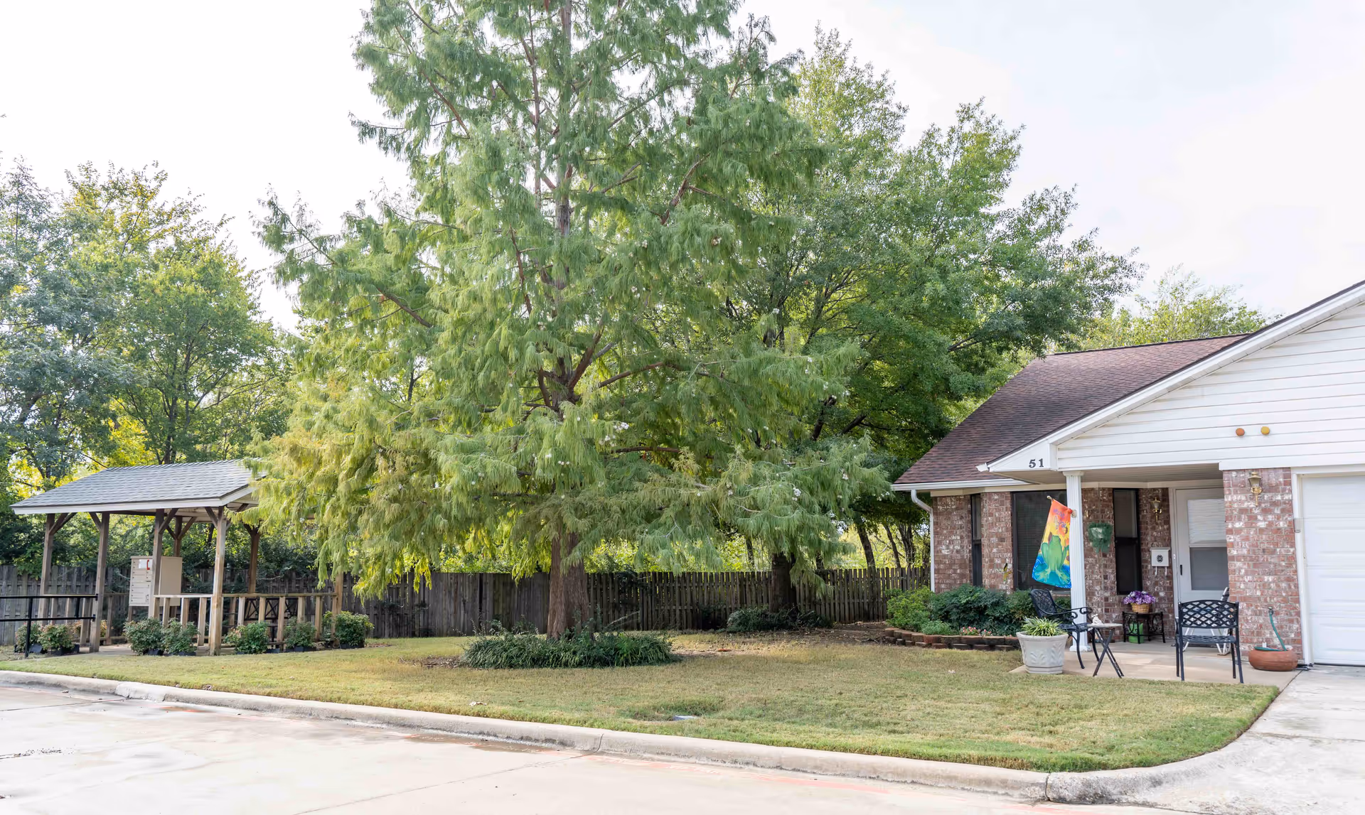 Single-story brick senior living residence with a front lawn, large trees, and porch seating.