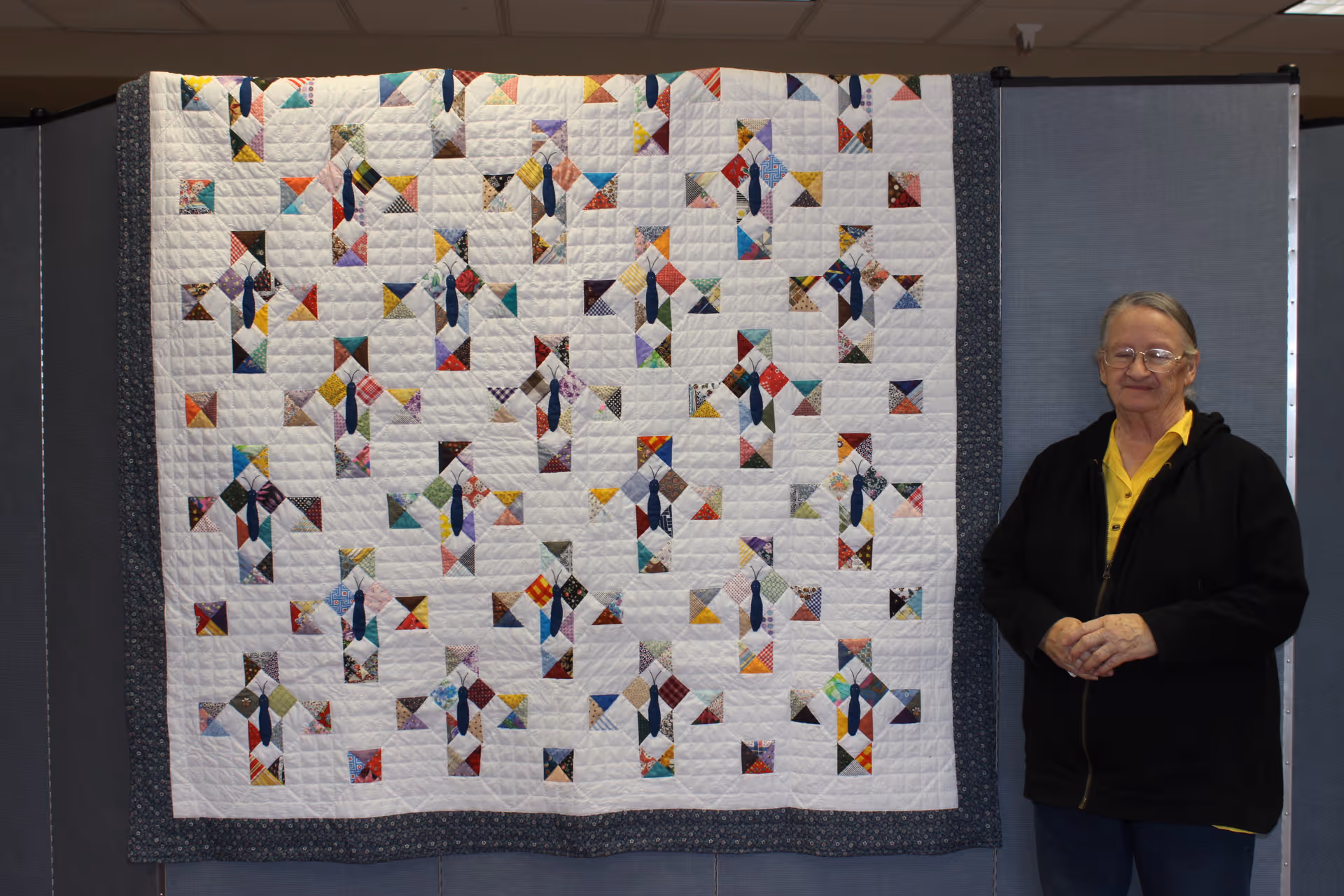 Elderly woman stands beside a colorful patchwork quilt displayed on a divider indoors.