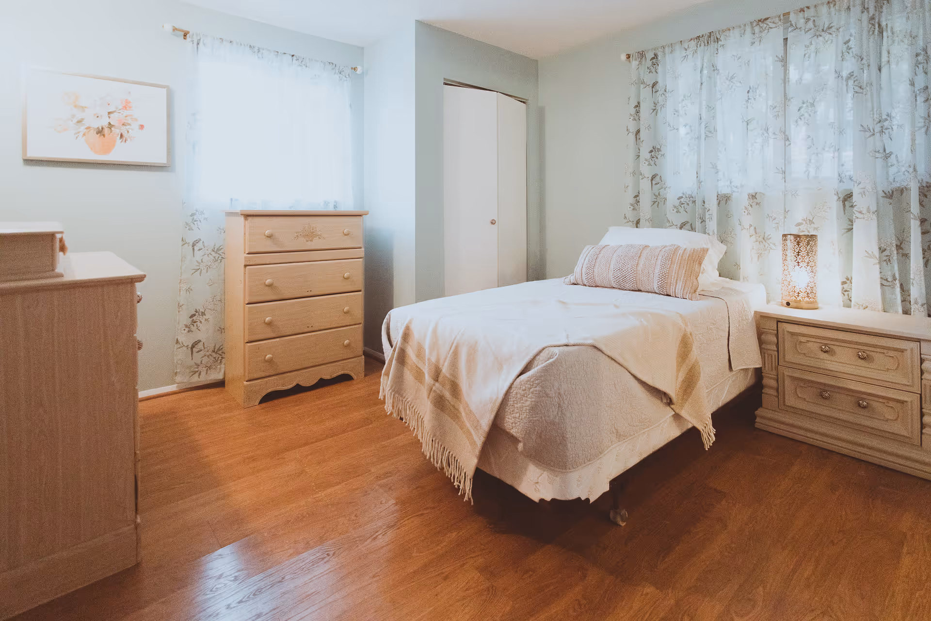 Light pastel bedroom with a bed, dresser, nightstands, and floral curtains.