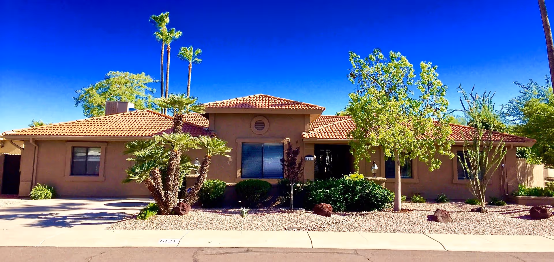 Single-story building with a tiled roof and beige exterior walls, surrounded by desert landscaping including palm trees, bushes, and rocks under a clear blue sky.